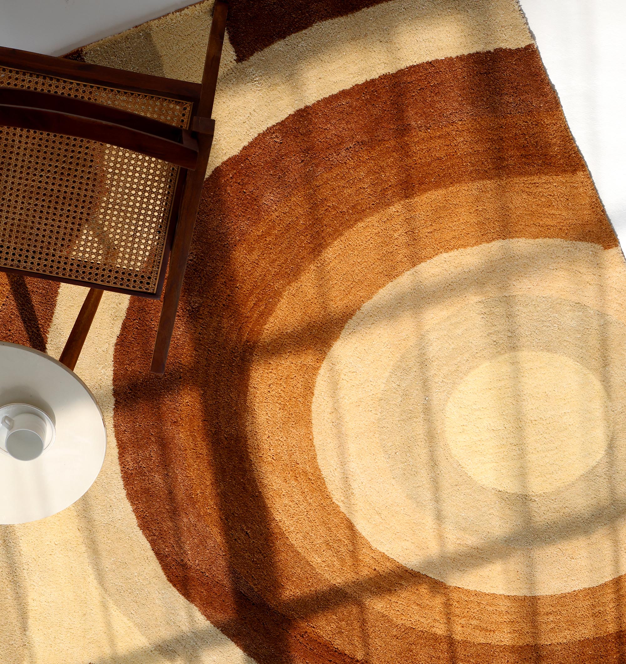 A wooden chair and a small white round table rest on the RAMSHA HOME Modern Beige & Brown Circular Shaggy Rug for Living Room (SG 053), with window shadows cast across the cozy scene.