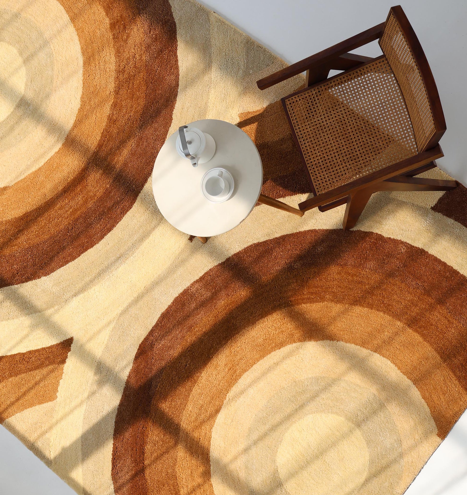 A top view of a round wooden chair, a small round table with a teapot and cup, and the RAMSHA HOME Modern Beige & Brown circular Shaggy Rug for Living Room - SG 053, all bathed in natural sunlight through window panes.