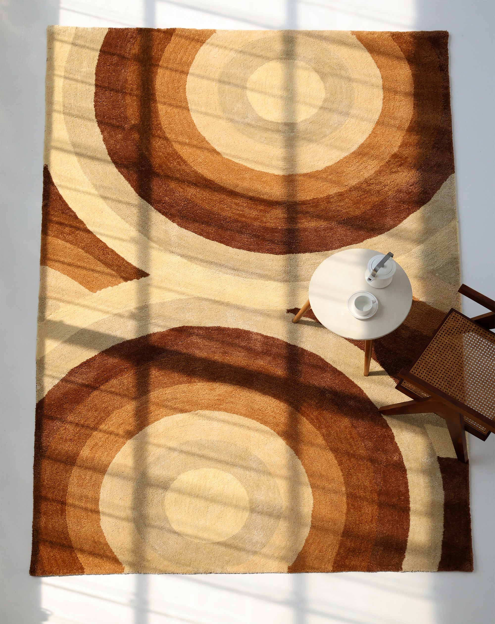 The RAMSHA HOME Modern Beige & Brown Circular Shaggy Rug for Living Room (SG 053) lies under a round table with a teapot and cup, while a wooden chair and sunlight create a cozy, inviting ambience on the soft rug.