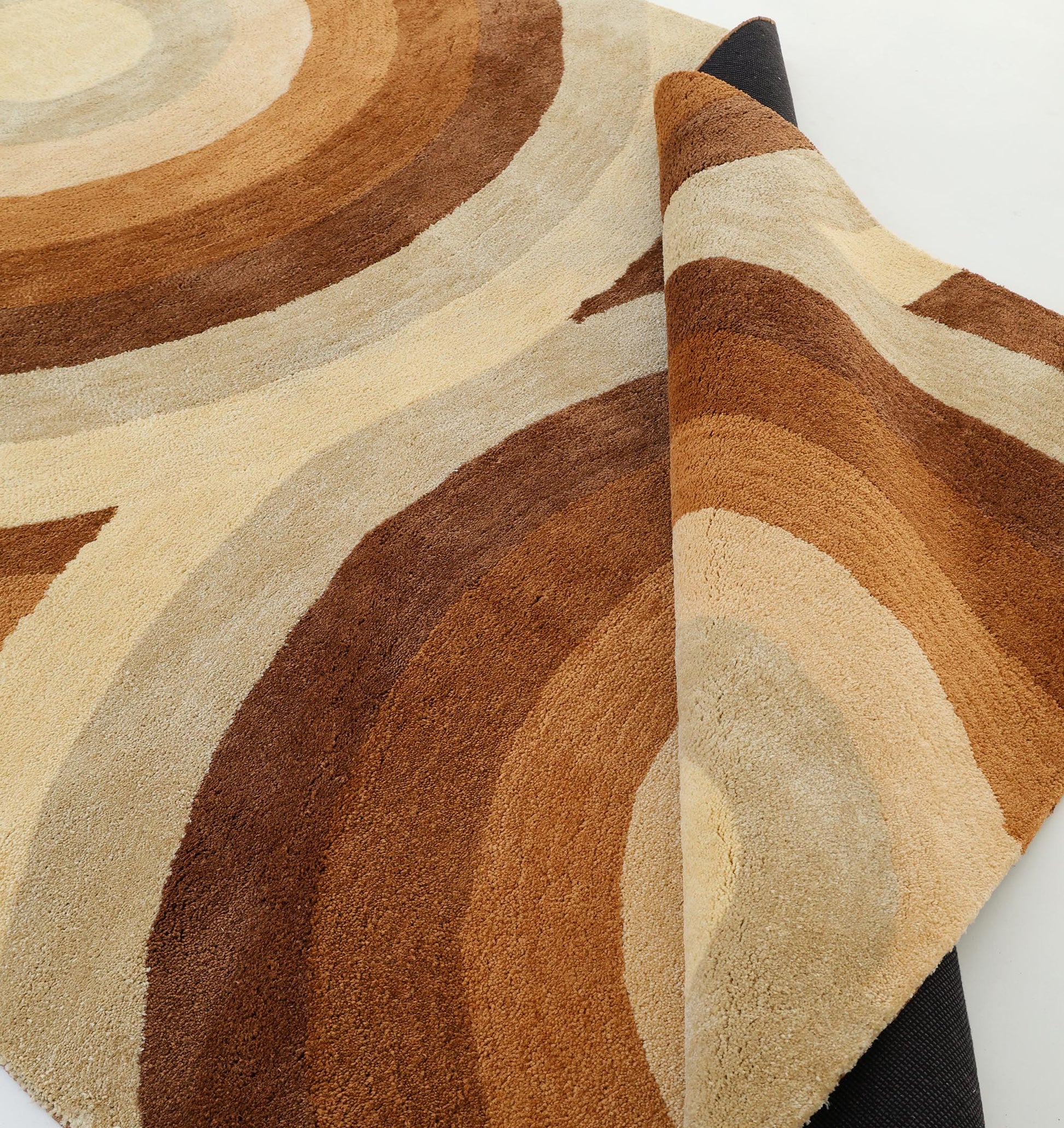 A close-up of the RAMSHA HOME Modern Beige & Brown circular Shaggy Rug for Living Room - SG 053, featuring curved bands in beige, tan, and brown. One corner is folded to show its dark backing.