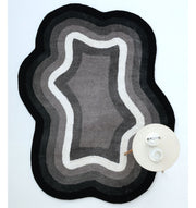 The RAMSHA HOME Modern Border Layered Irregular Soft Shaggy Carpet - SG 054, in black, gray, and white with wavy edges, rests on a white floor. A round table with a teapot and cup is placed on the carpet’s lower right corner.
