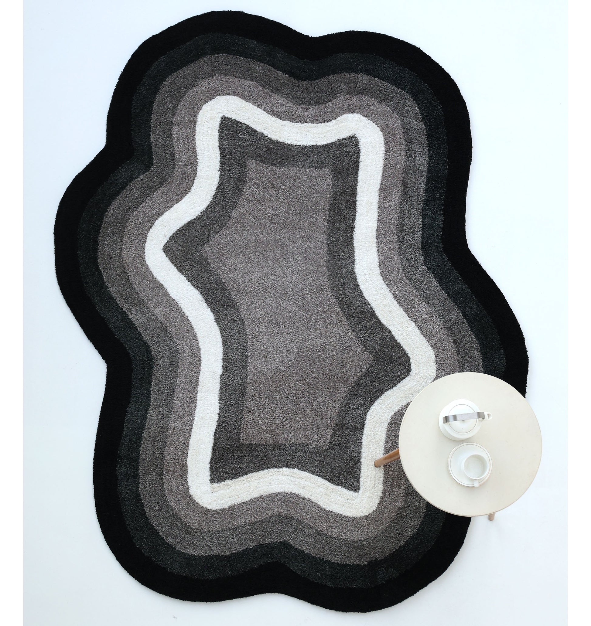 The RAMSHA HOME Modern Border Layered Irregular Soft Shaggy Carpet - SG 054, in black, gray, and white with wavy edges, rests on a white floor. A round table with a teapot and cup is placed on the carpet’s lower right corner.