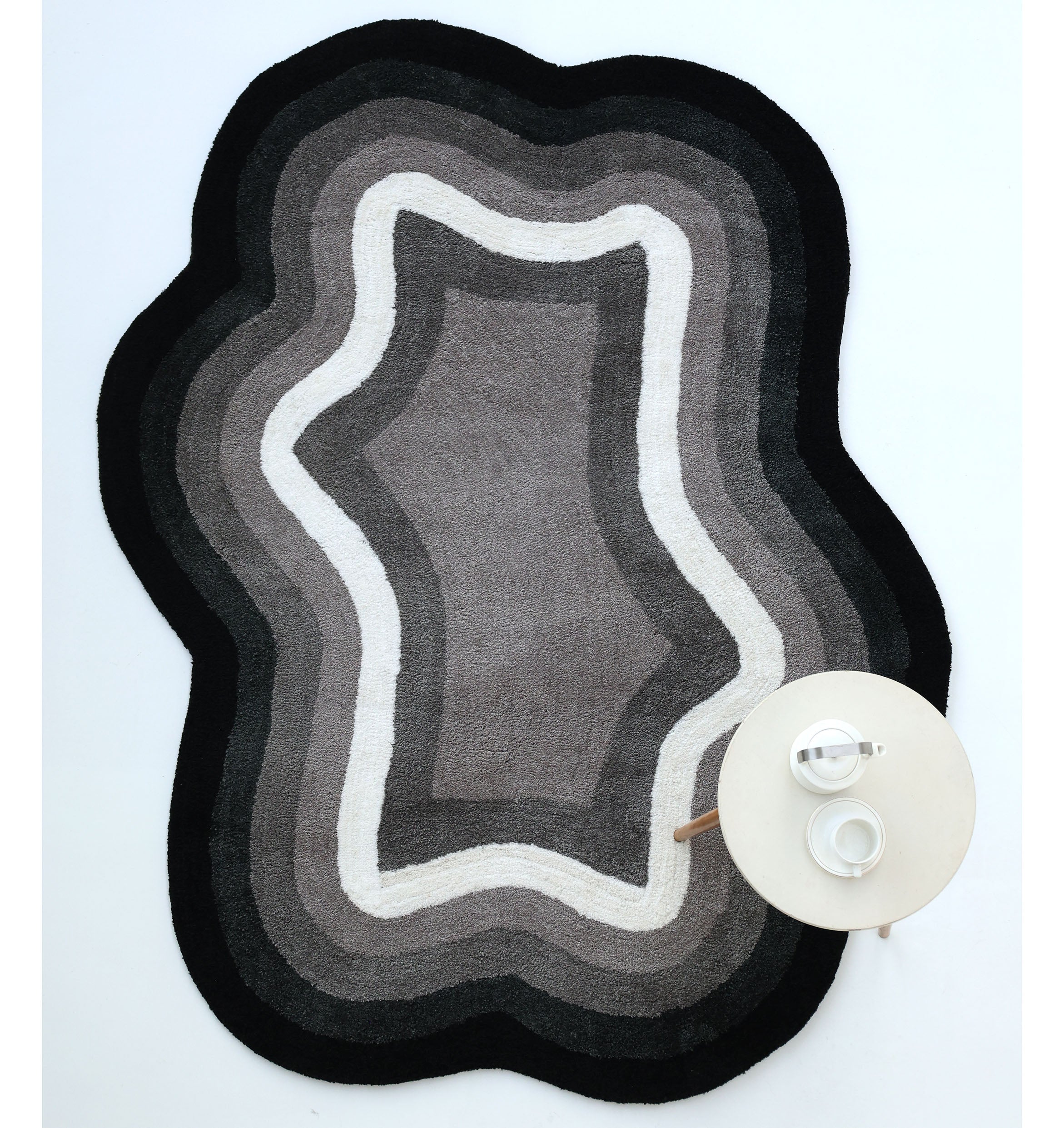 The RAMSHA HOME Modern Border Layered Irregular Soft Shaggy Carpet - SG 054, in black, gray, and white with wavy edges, rests on a white floor. A round table with a teapot and cup is placed on the carpet’s lower right corner.