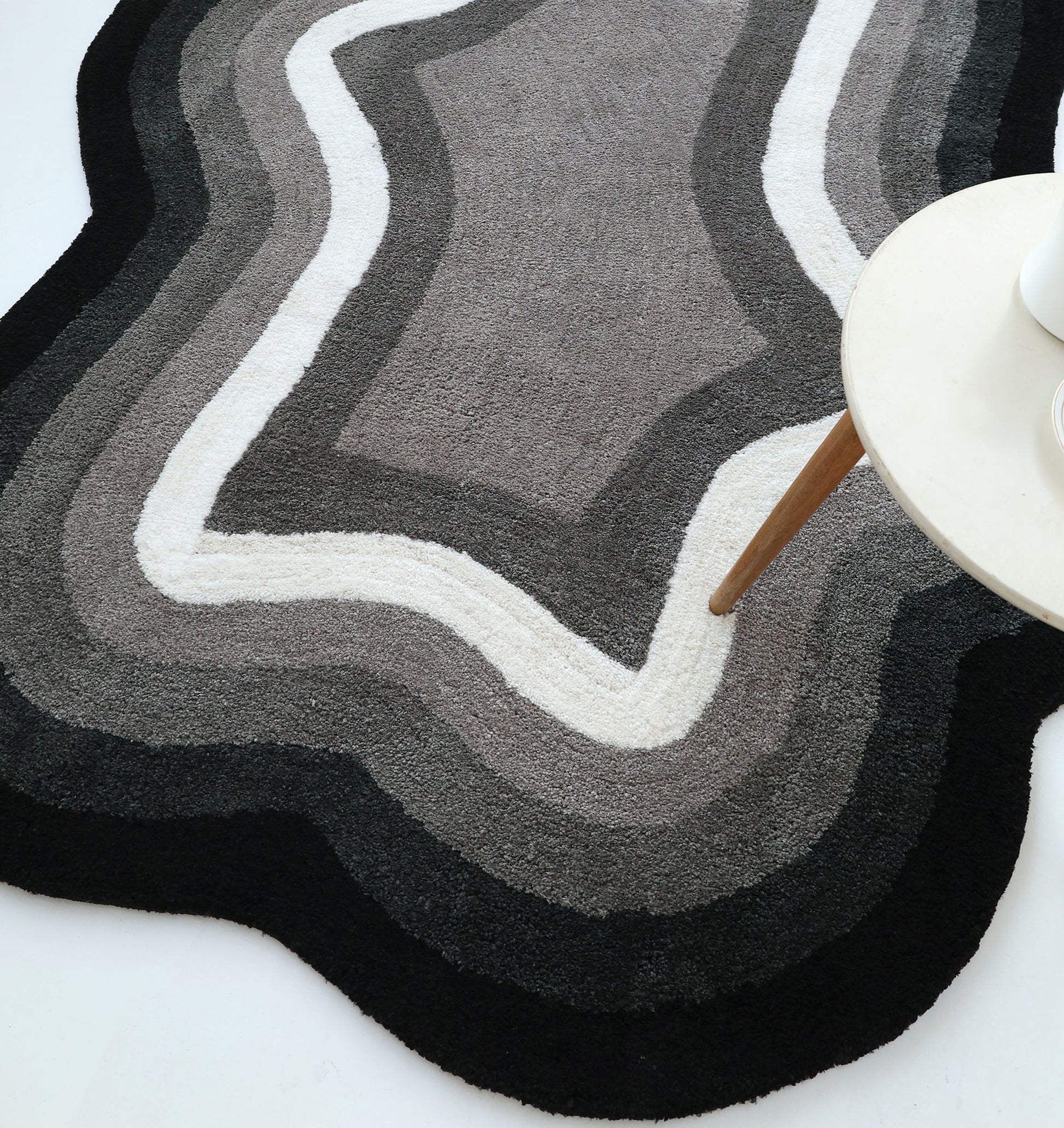 The RAMSHA HOME Modern Border Layered Irregular Soft Shaggy Carpet - SG 054, featuring wavy black, gray, and white layers, lies on a white floor beside a round light table with wooden legs.