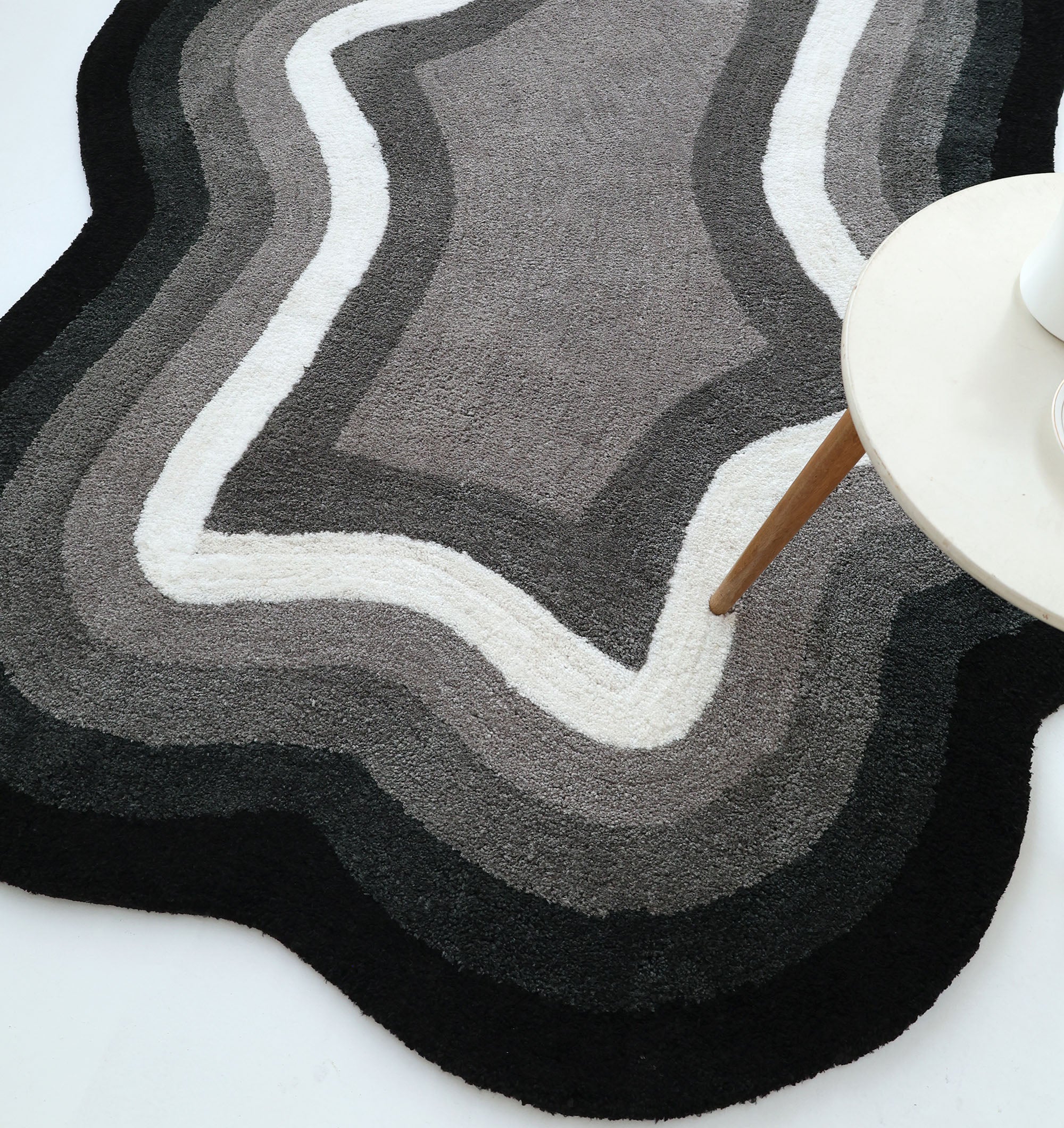 The RAMSHA HOME Modern Border Layered Irregular Soft Shaggy Carpet - SG 054, featuring wavy black, gray, and white layers, lies on a white floor beside a round light table with wooden legs.
