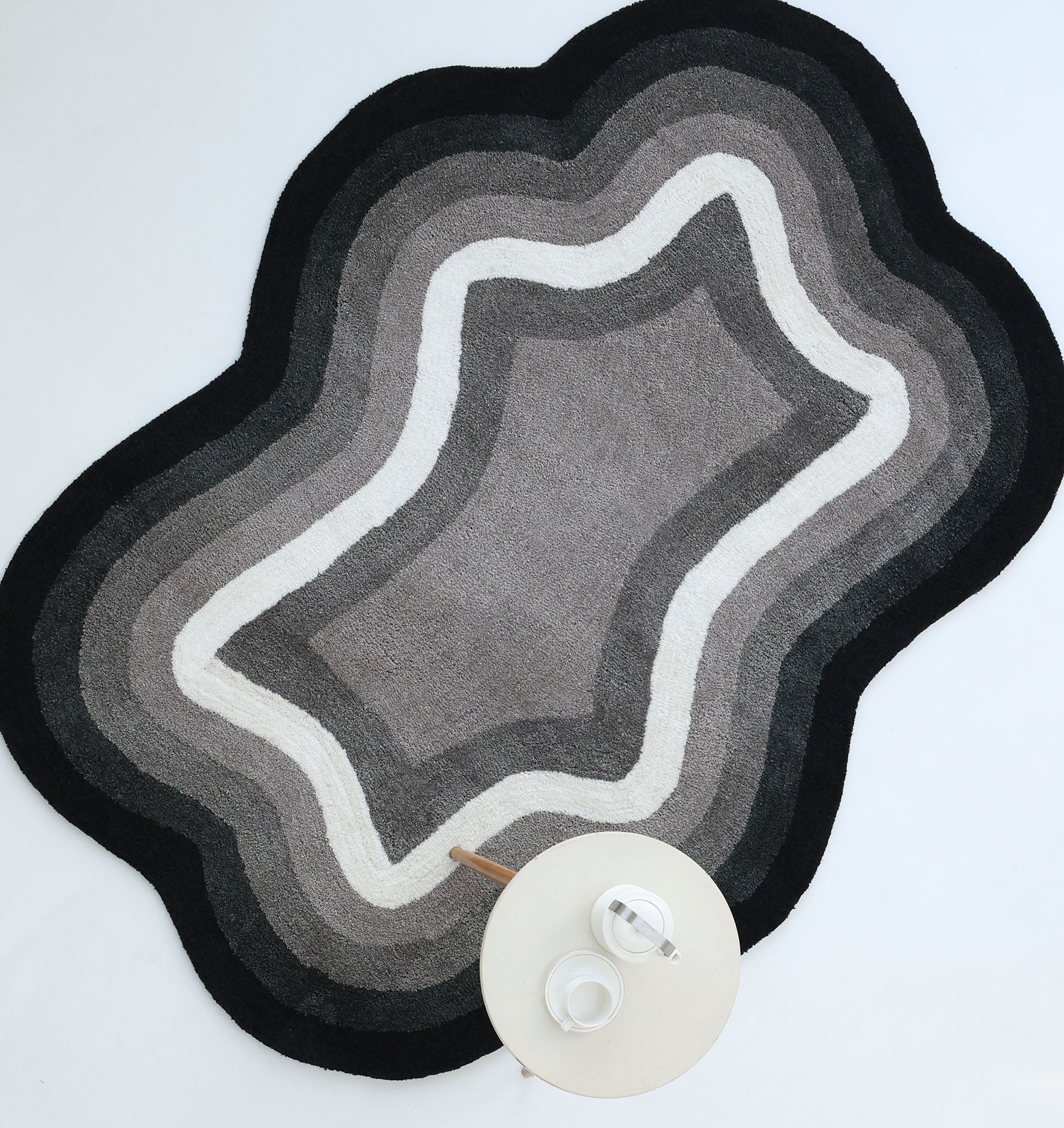 The RAMSHA HOME Modern Border Layered Irregular Soft Shaggy Carpet - SG 054, in wavy shades of gray, white, and black, lies on the floor beside a round white side table with a teapot and cup.