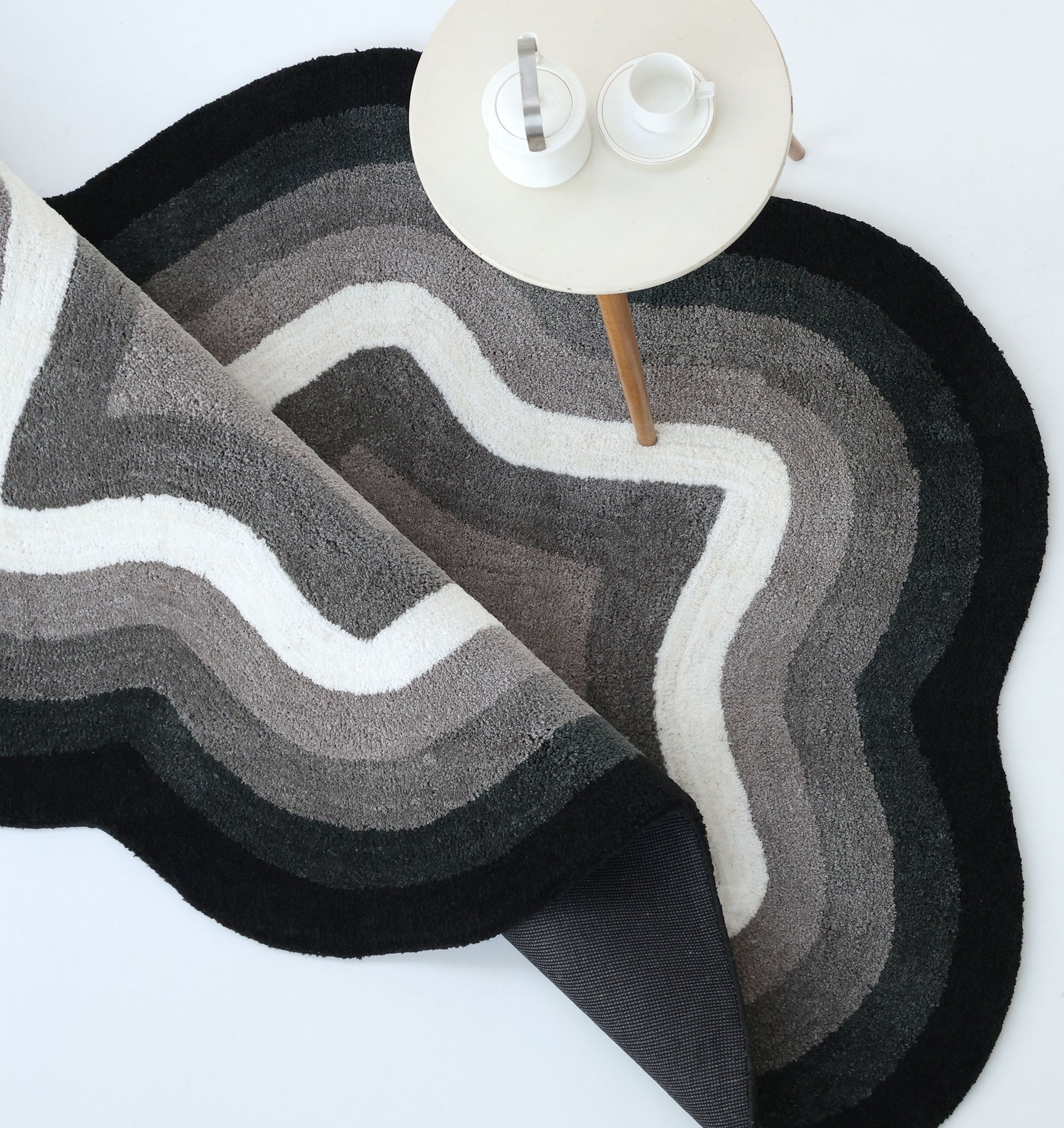The RAMSHA HOME Modern Border Layered Irregular Soft Shaggy Carpet - SG 054 in black, gray, and white sits partially rolled up beside a small round table with a teapot, sugar jar, and cup on a light-colored surface.