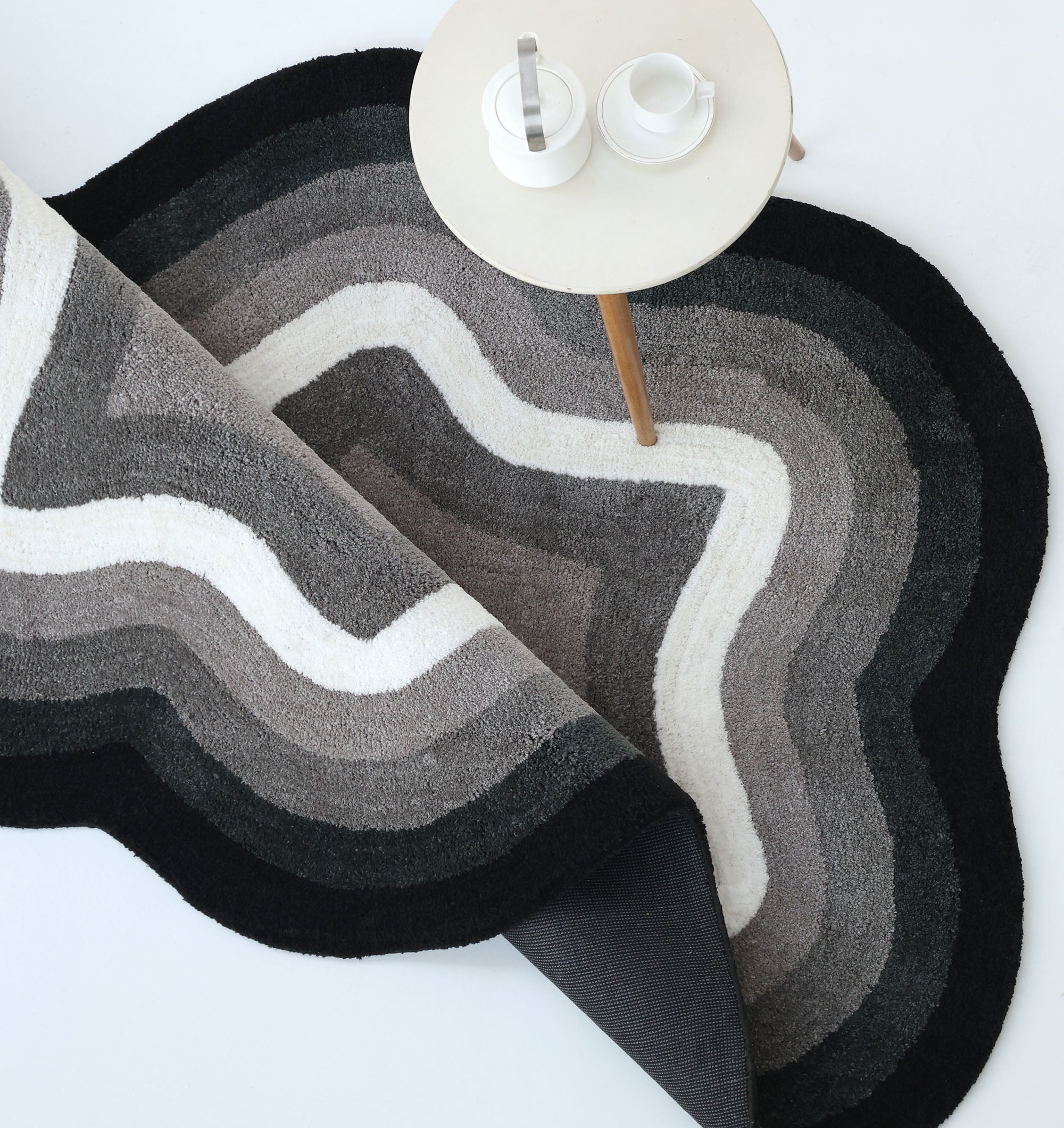The RAMSHA HOME Modern Border Layered Irregular Soft Shaggy Carpet - SG 054 in black, gray, and white sits partially rolled up beside a small round table with a teapot, sugar jar, and cup on a light-colored surface.
