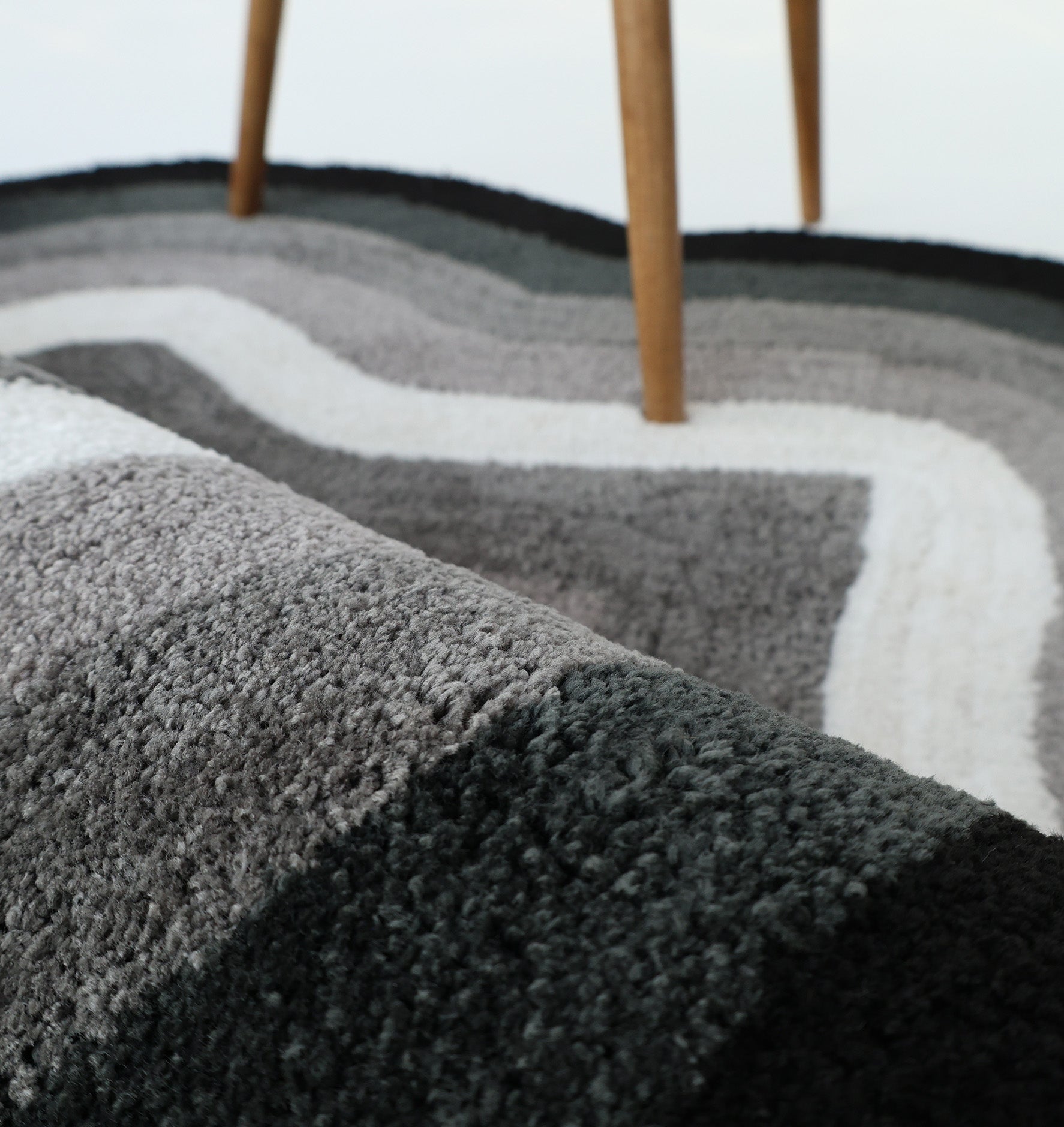 A close-up of the RAMSHA HOME Modern Border Layered Irregular Soft Shaggy Carpet - SG 054 in gray, white, and black wavy patterns, with wooden furniture legs visible in the background.