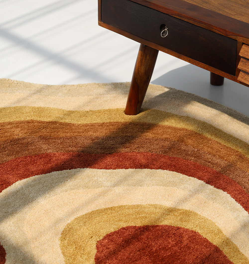 A close-up of a wooden table leg and drawer above the RAMSHA HOME Modern Layered Border Irregular Soft Shaggy Carpet - SG 055, featuring beige, mustard yellow, and rust wavy patterns—perfect for cozy home décor in natural sunlight.