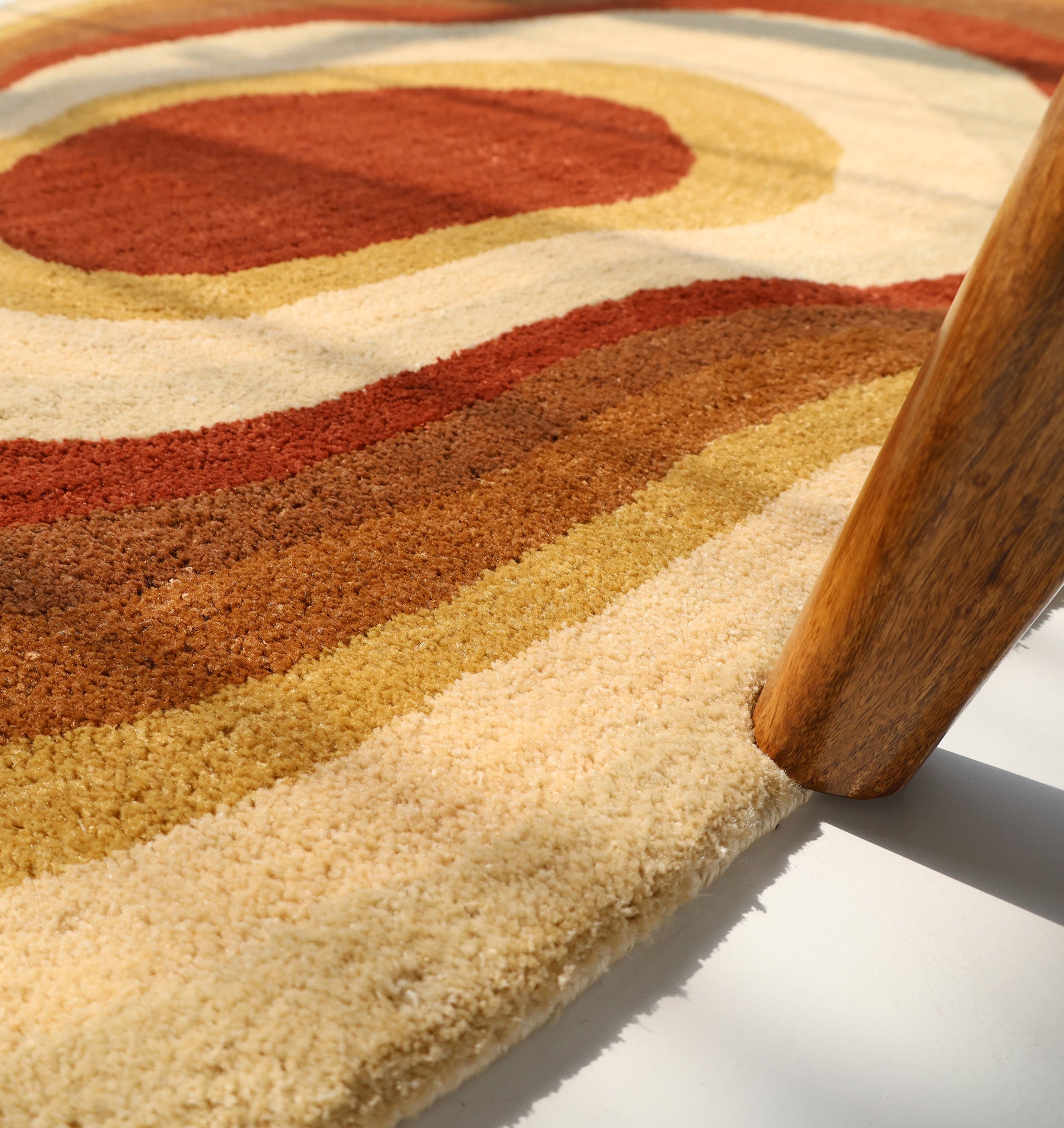A close-up of the RAMSHA HOME Modern Layered Border Irregular Soft Shaggy Carpet - SG 055 shows its cream, brown, and rust concentric circles. A wooden furniture leg rests on the plush rug, adding an inviting touch to cozy home décor.