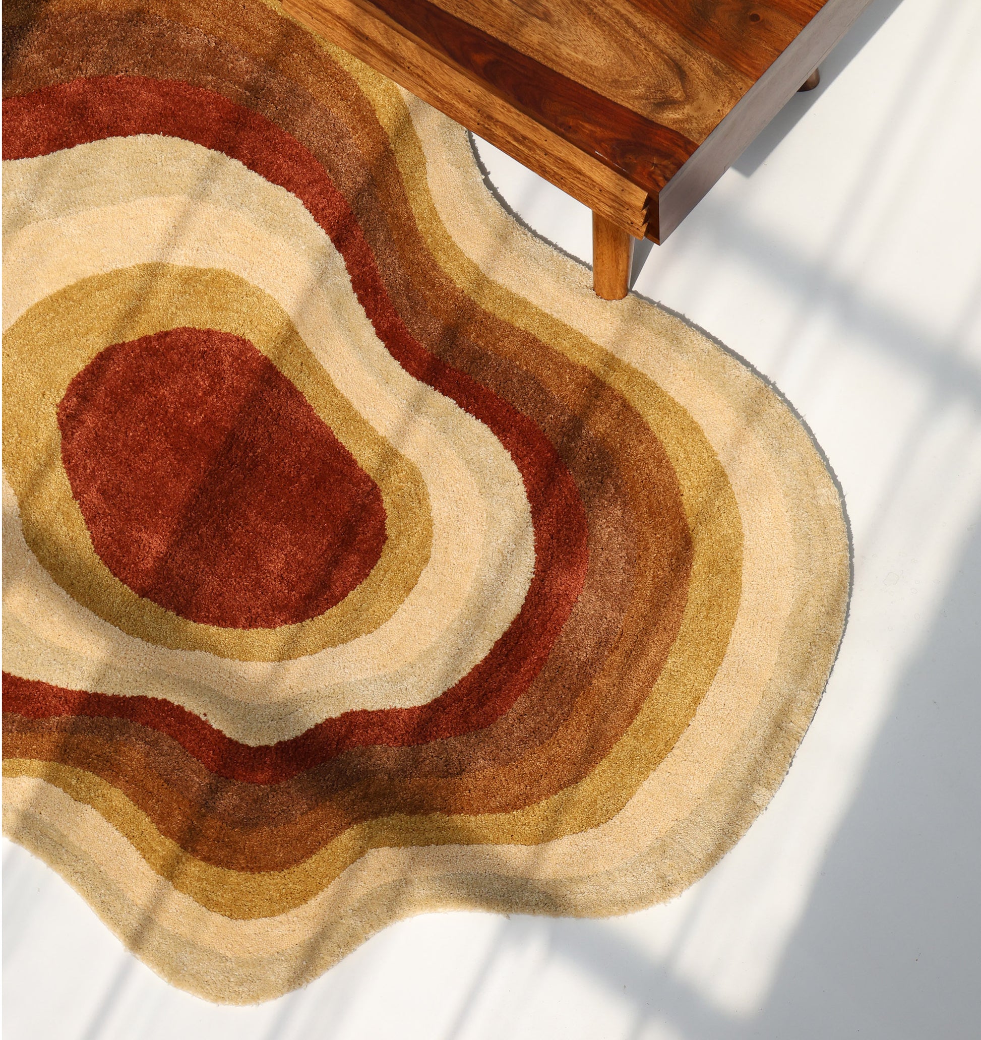 The RAMSHA HOME Modern Layered Border Irregular Soft Shaggy Carpet - SG 055, featuring wavy, multicolored shapes in brown, beige, and rust, lies on a white floor beside a wooden table with sunlight casting gentle shadows.
