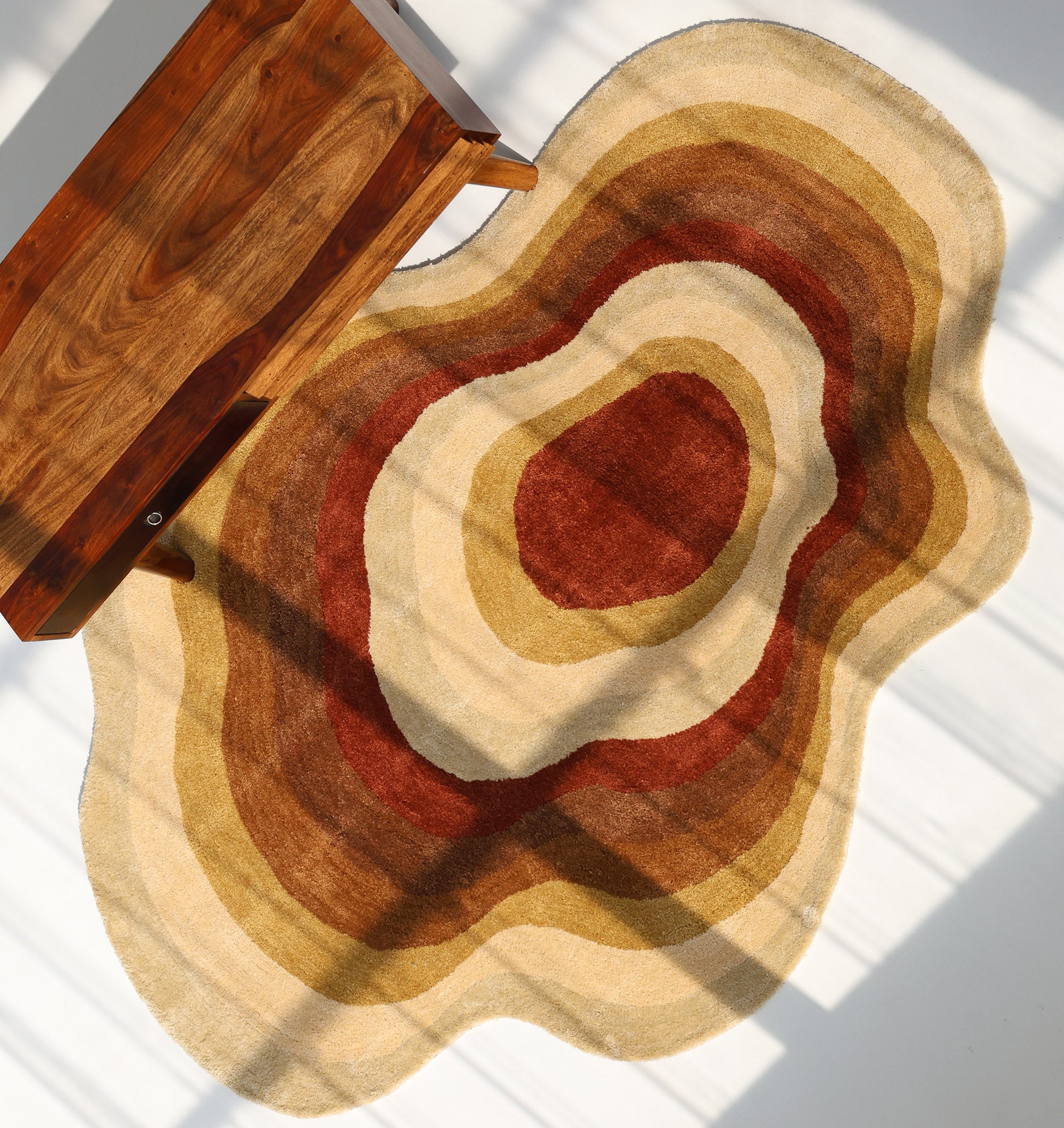 The RAMSHA HOME Modern Layered Border Irregular Soft Shaggy Carpet - SG 055, featuring abstract earth-toned rings in brown, beige, and rust, lies on a white floor near a wooden bench, with windowpane shadows creating a cozy atmosphere.