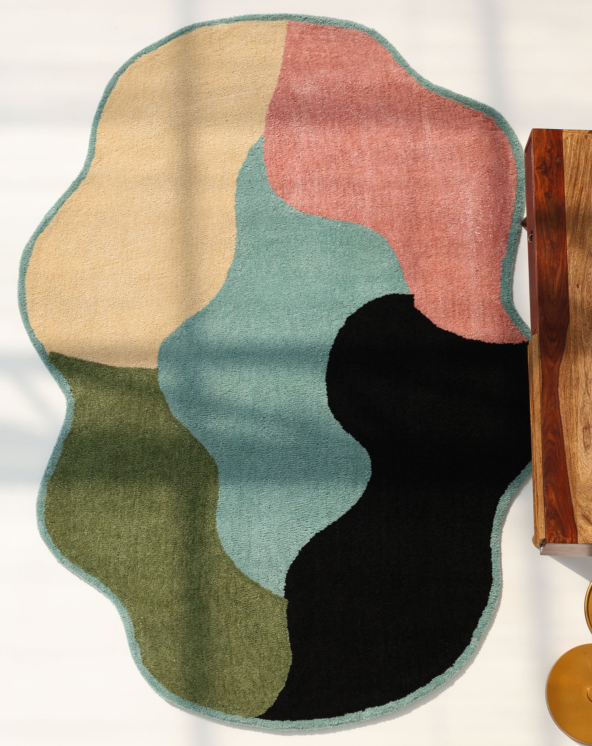 The RAMSHA HOME Modern multicolor Irregular Soft Shaggy Carpet - SG 056 showcases abstract wavy patches in vibrant hues with a teal border, partly under a wooden table, sunlight enhancing its cozy and stylish appeal.