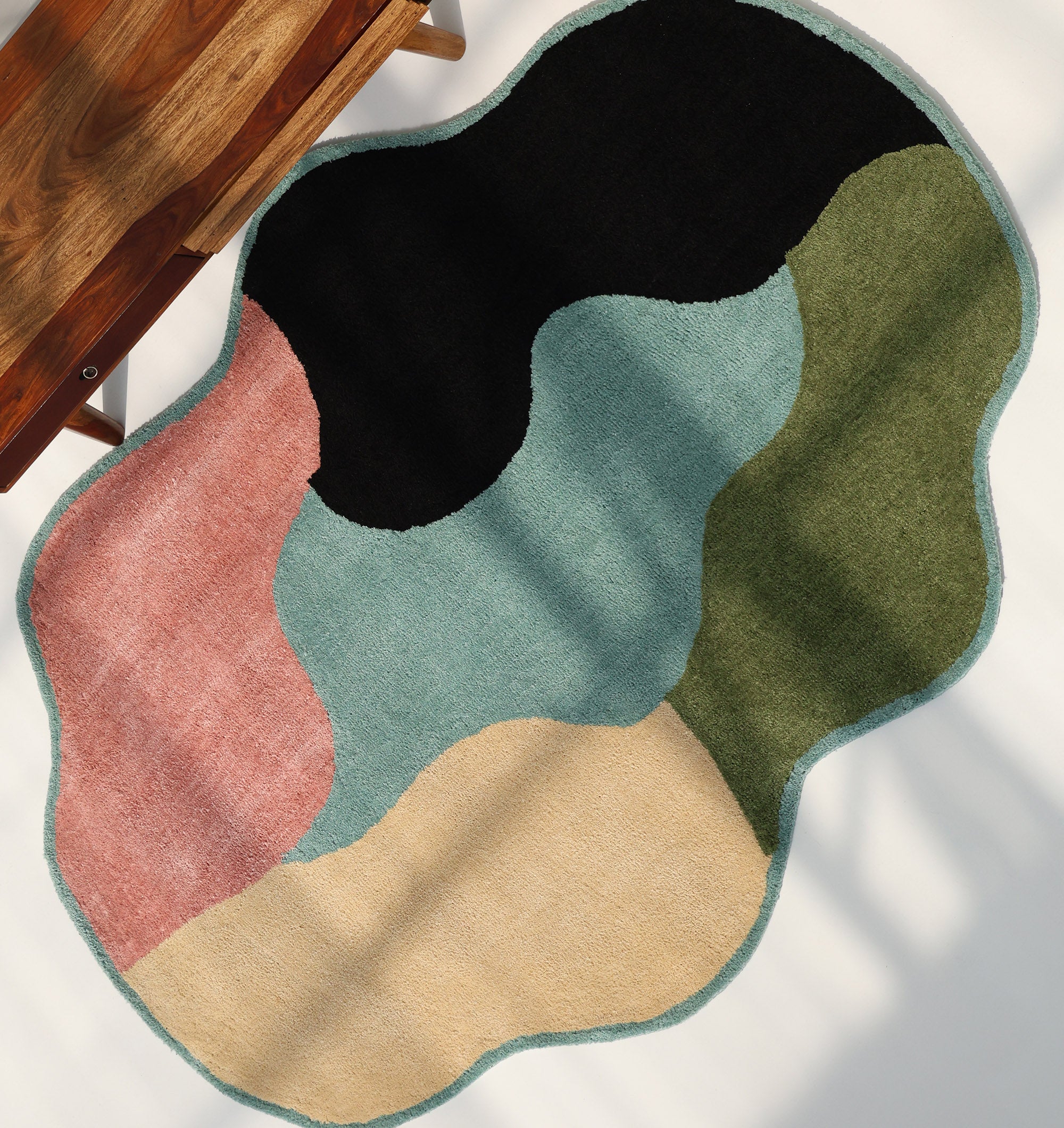 The RAMSHA HOME Modern Multicolor Irregular Soft Shaggy Carpet - SG 056, with wavy sections in black, green, blue, pink, and beige, adds cozy style next to a wooden bench on a white surface with shadow patterns.