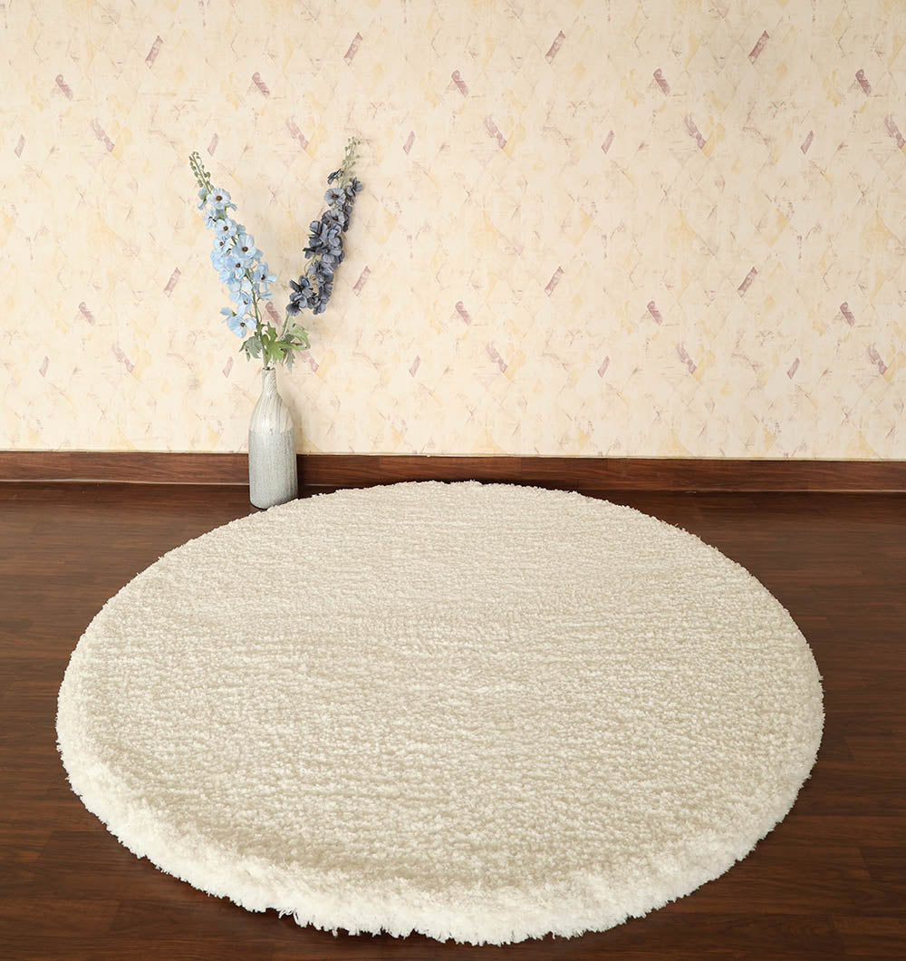 The RAMSHA HOME Round Soft White Shaggy Rug - SG 06 sits on a dark wooden floor with textured beige wallpaper; a tall glass vase of blue and purple flowers nearby enhances the elegant scene.