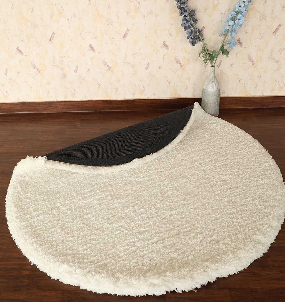 The RAMSHA HOME Round Soft White Round Shaped Shaggy Rug - SG 06, with a black non-slip underside, is displayed on a wooden floor near a wall featuring a vase of blue and purple flowers.