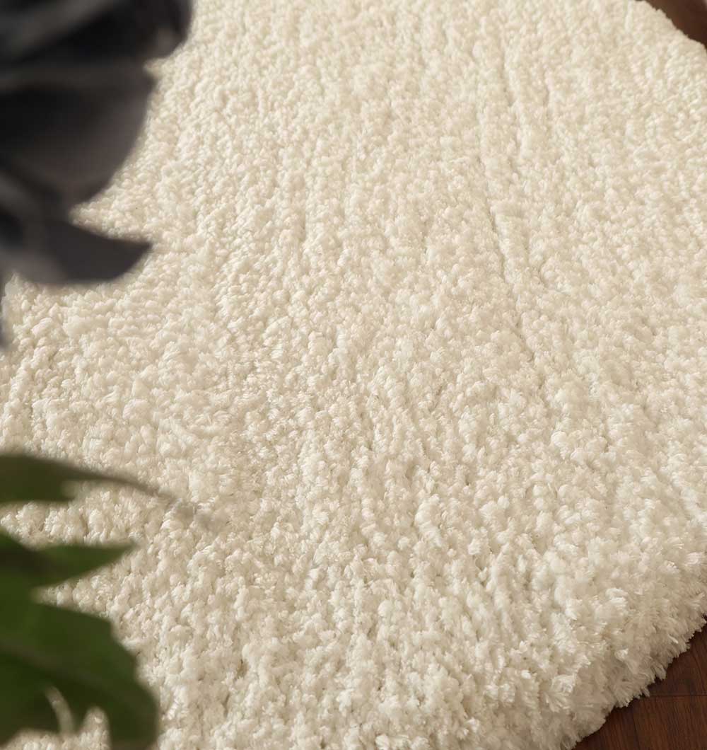 Close-up of the RAMSHA HOME Round Soft White Shaggy Rug - SG 06 on a wooden floor, with blurred green leaves and a dark flower in the upper left corner.