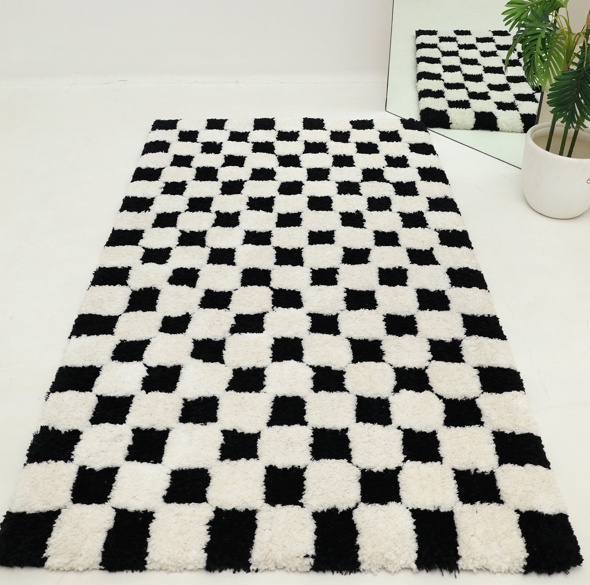 The RAMSHA HOME Black & White Checkered Shaggy Rug 115x230 cm (SG-069), featuring a classic checkerboard pattern, is spread out on a white floor. Its reflection and a potted plant can be seen in a nearby mirror at the corner.