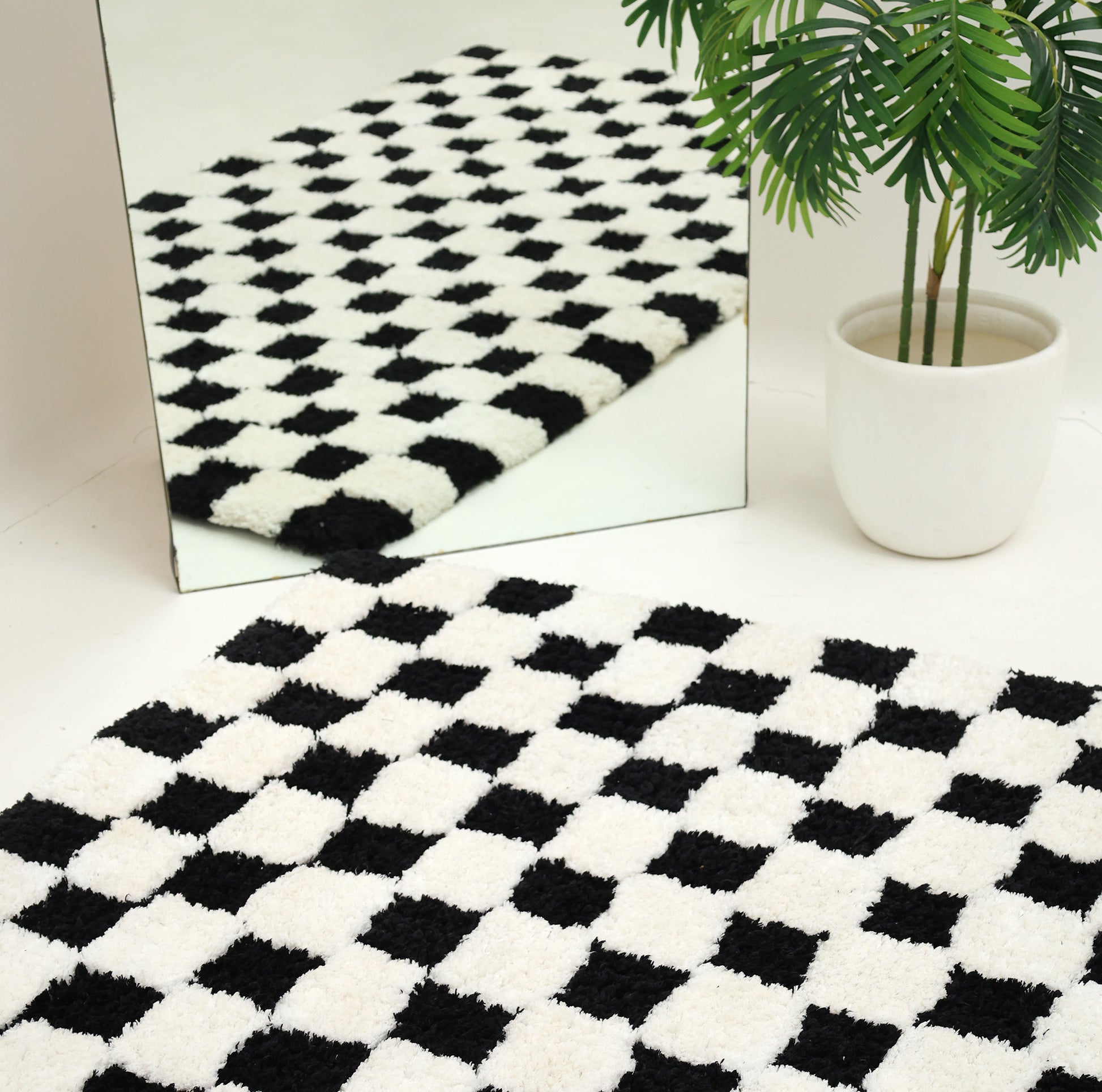 A RAMSHA HOME Black & White Checkered Shaggy Rug (115x230 cm, SG-069) is laid on the floor beside a large mirror and a potted green plant with broad leaves. The rug’s bold checkerboard pattern is reflected in the mirror.