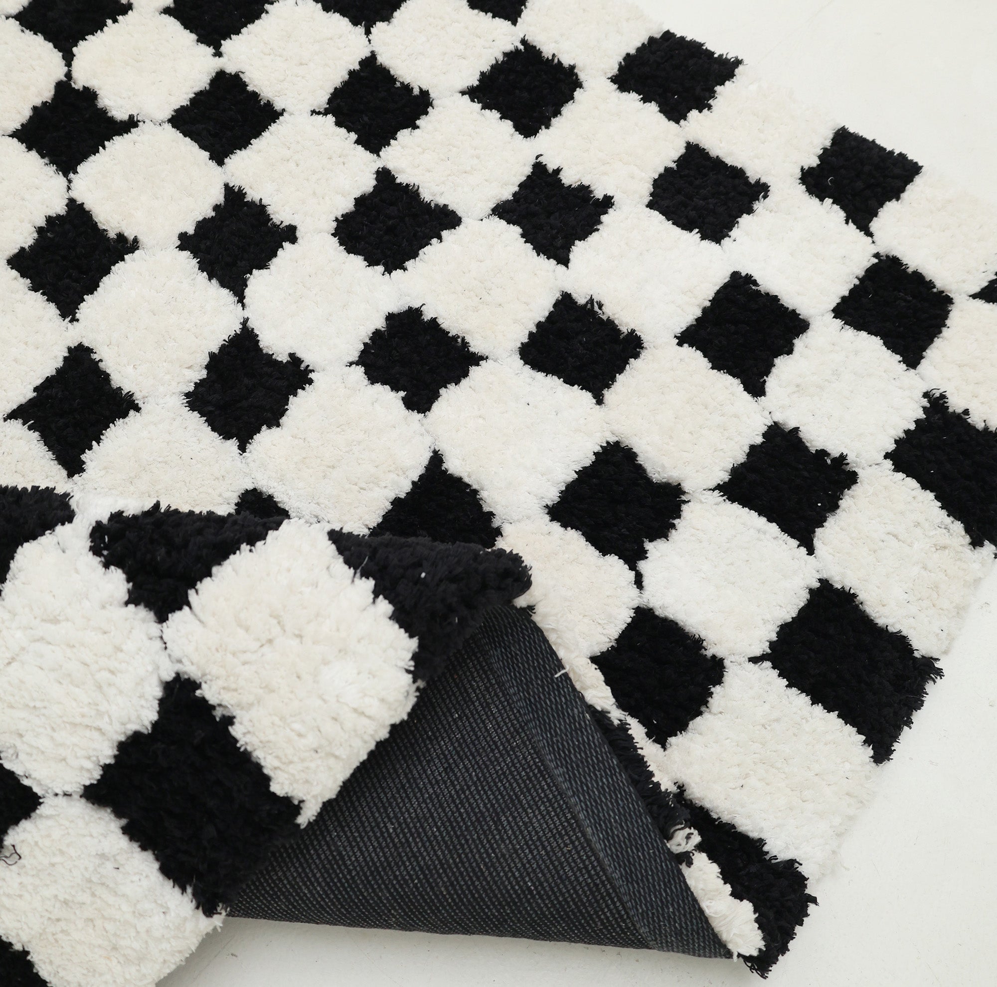A close-up of the RAMSHA HOME Black & White Checkered Shaggy Rug 115x230 cm (SG-069) highlights its bold black and white squares. One corner is lifted to reveal a dark non-slip backing, while the soft, textured surface exudes comfort.