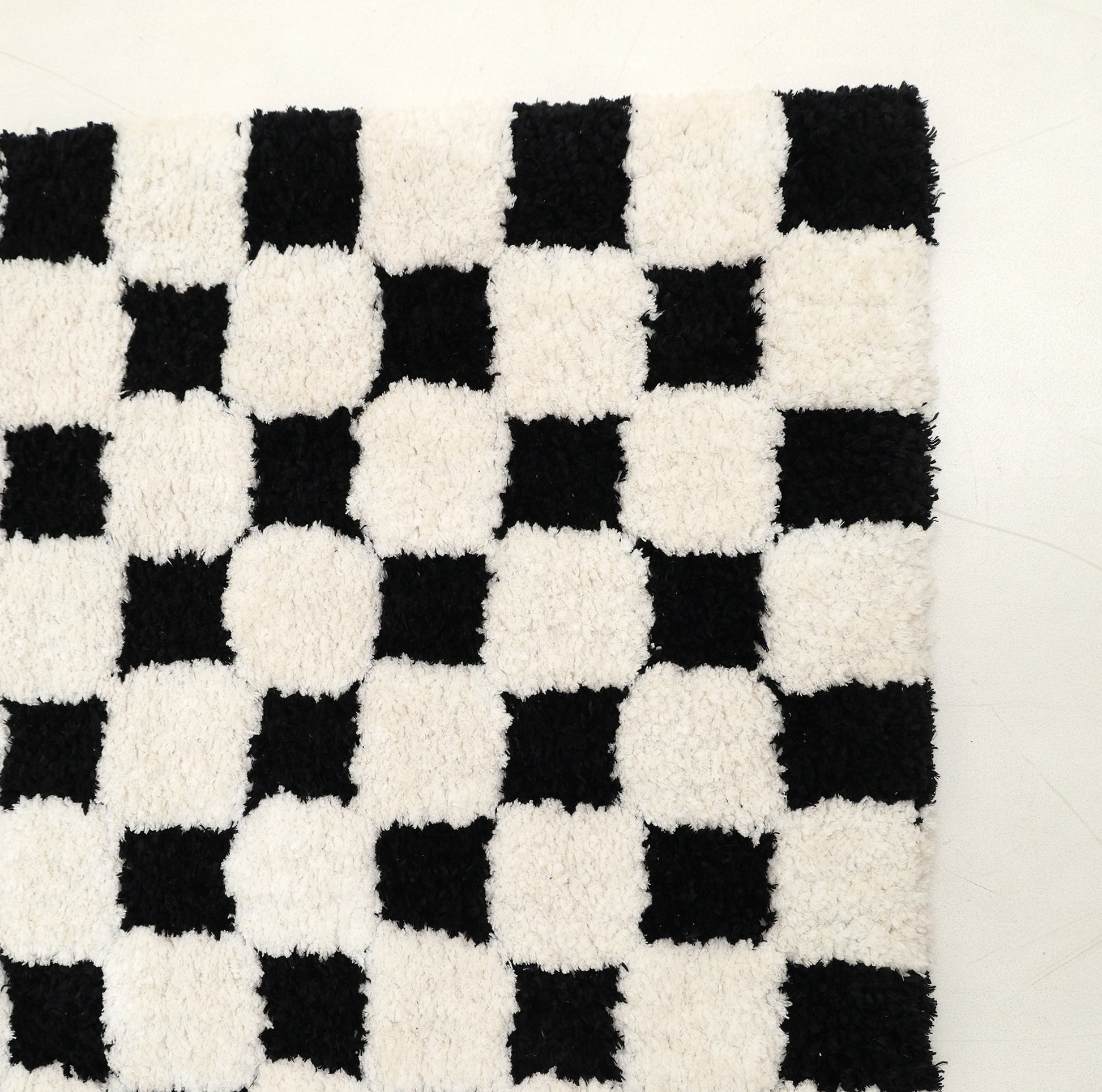 The RAMSHA HOME Black & White Checkered Shaggy Rug 115x230 cm (SG-069) showcases an eye-catching geometric design with alternating black and white squares. Its bold checkerboard pattern stands out beautifully against a plain, light-colored floor.