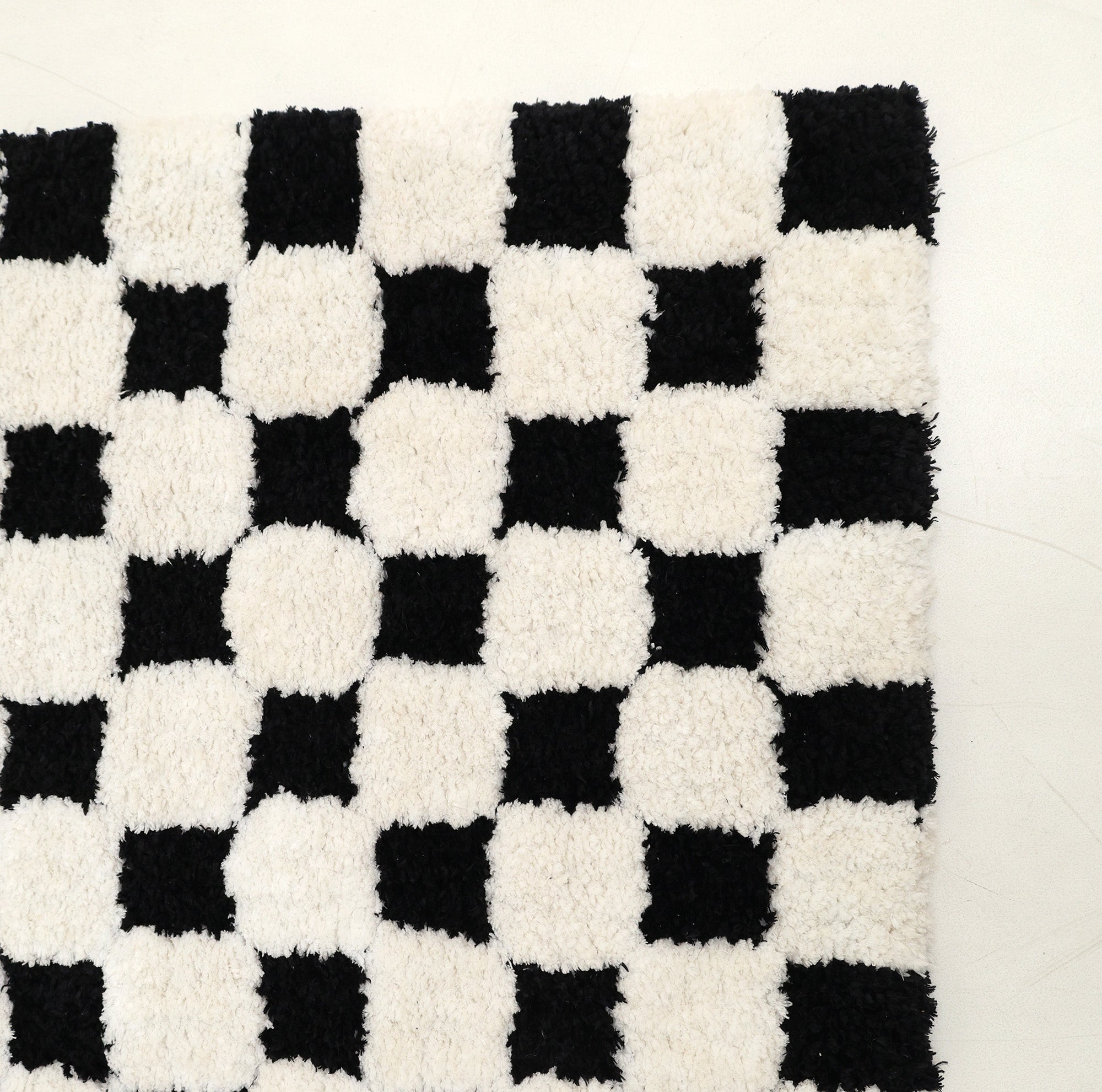 The RAMSHA HOME Black & White Checkered Shaggy Rug 115x230 cm (SG-069) showcases an eye-catching geometric design with alternating black and white squares. Its bold checkerboard pattern stands out beautifully against a plain, light-colored floor.