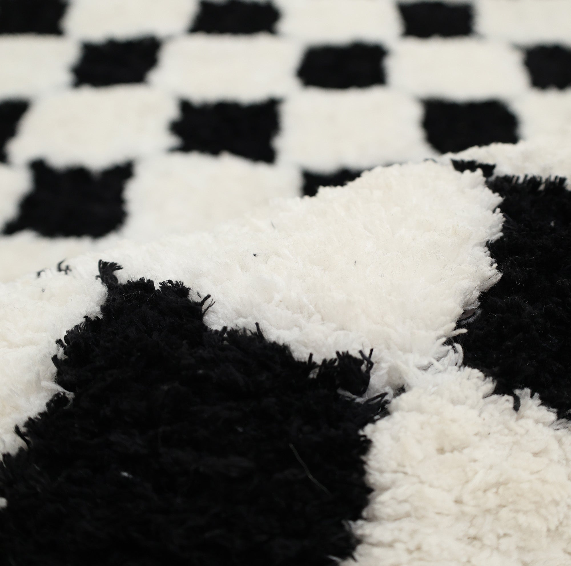 A close-up of the RAMSHA HOME Black & White Checkered Shaggy Rug (115x230 cm, SG-069) reveals its soft, textured fibers and slightly rumpled surface in the foreground.