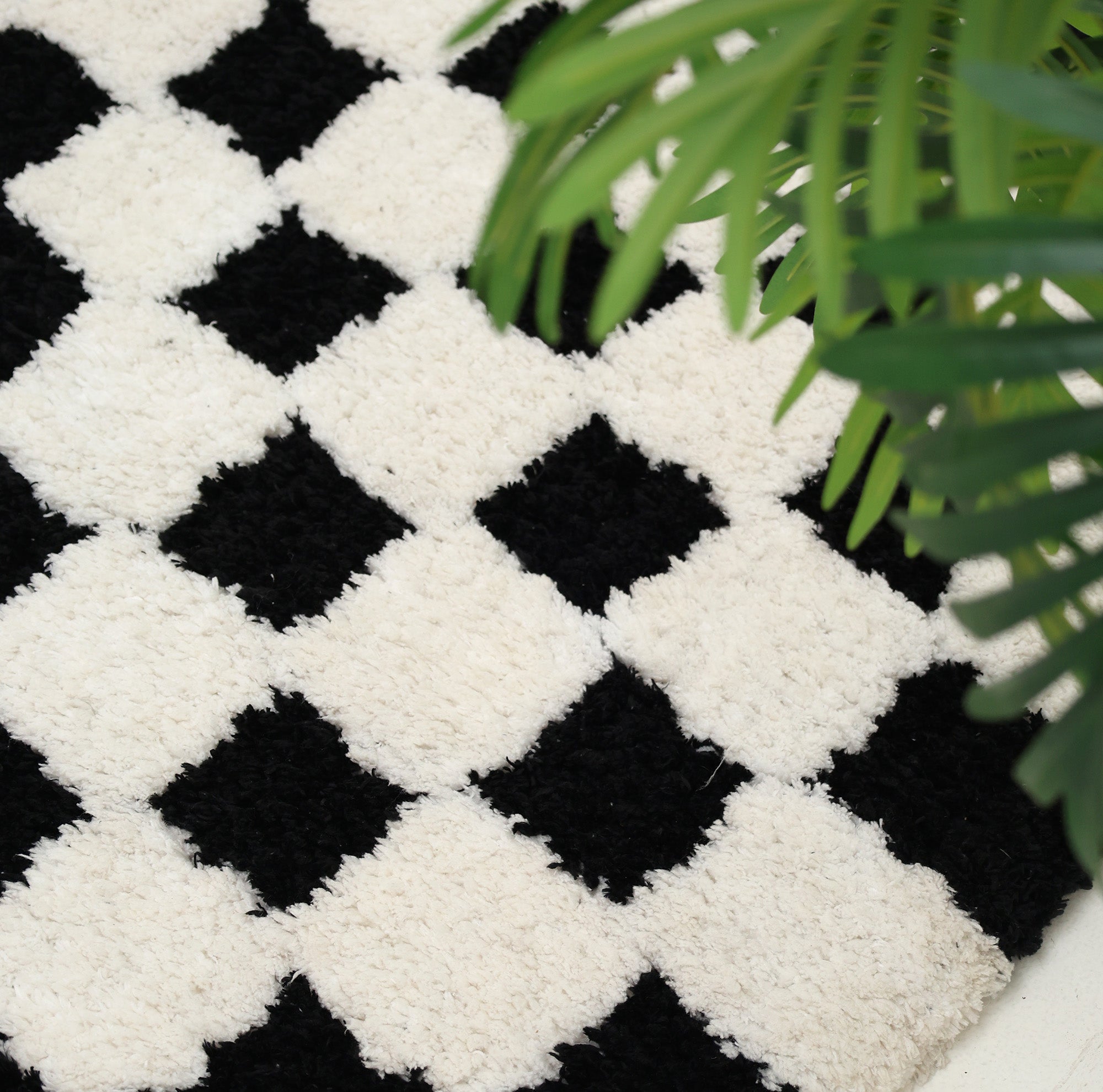 A close-up of the RAMSHA HOME Black & White Checkered Shaggy Rug 115x230 cm (SG-069), featuring a bold geometric diamond pattern, partially covered by green leaves from a nearby plant.