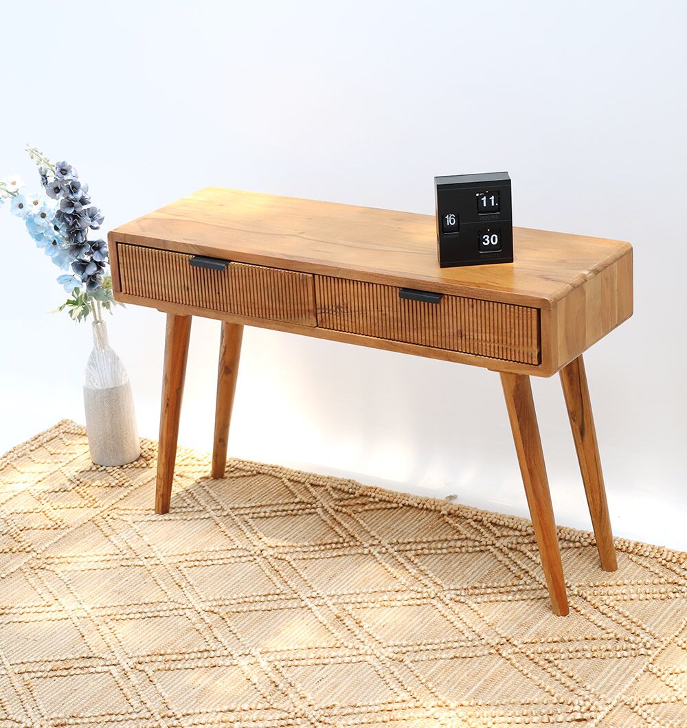 A compact workspace features the RAMSHA HOME Wooden Sideboard Cabinet with Storage – Solid Acacia, holding a digital clock on top and blue and white flowers in a vase beside it, all set against a white wall on a patterned rug.