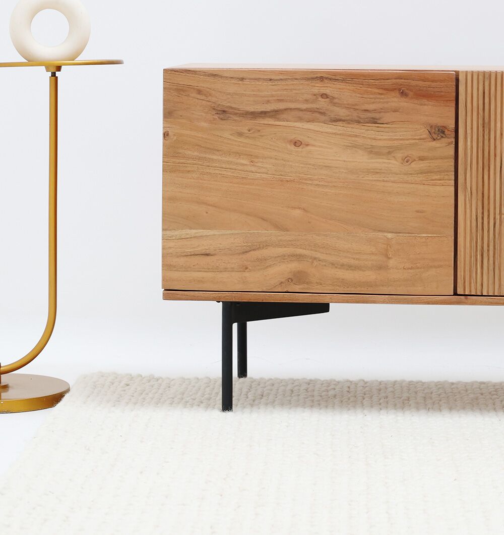 A minimal scene with the corner of the RAMSHA HOME Wooden TV Cabinet with Storage – Solid Acacia & Metal Base, a gold side table holding a round white decor object, and a white textured rug on the floor.