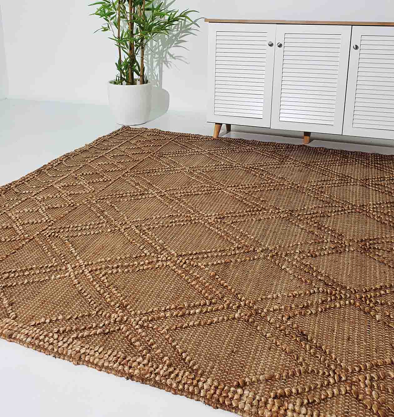 The RAMSHA HOME Floor Woven Living Room Rug SR-019, a large rectangular jute rug in natural color with a geometric diamond pattern, is displayed on a white floor next to a white slatted cabinet and a green potted plant.