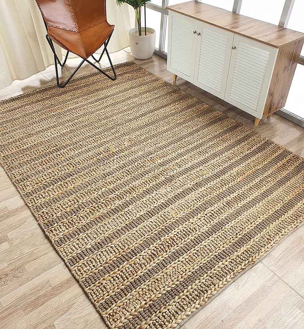 The RAMSHA HOME Jute Woven Home Decor Rug Natural Color SR-032, featuring alternating light and dark stripes, lies on a wooden floor beside a small cabinet and brown leather chair near sunlit windows.