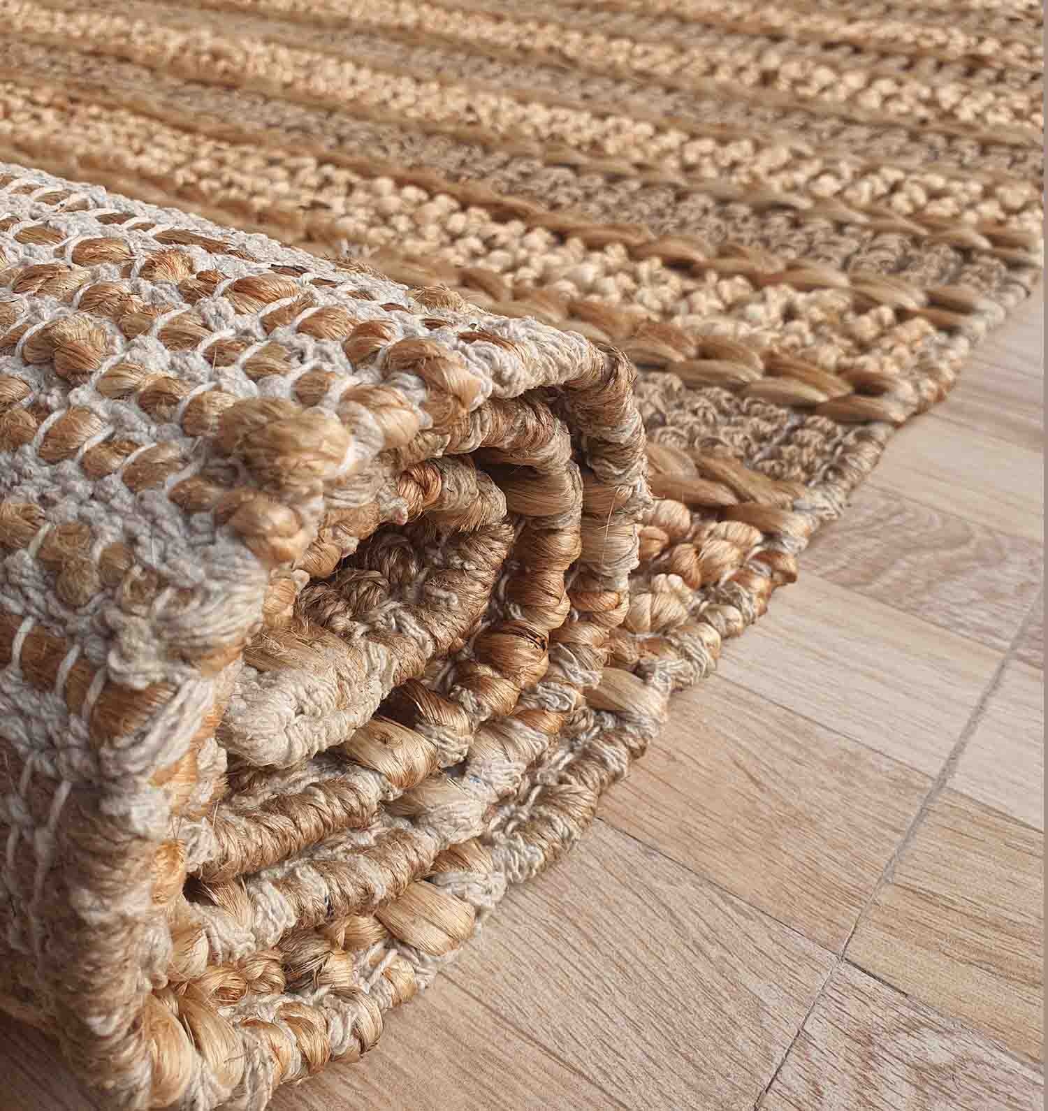 A close-up of the RAMSHA HOME Jute Woven Home Decor Rug Natural Color SR-032 shows its textured beige and brown weave, rolled up on a light wooden floor, highlighting the natural jute patterns.