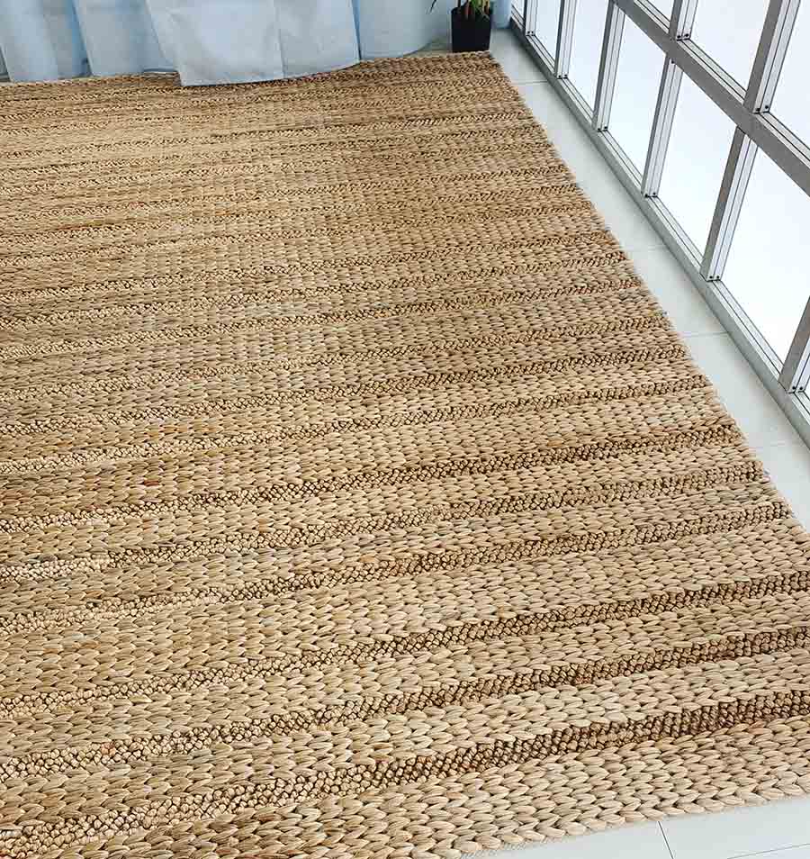 The RAMSHA HOME Natural Color Hand Woven Jute Rug SR-043, with its braided texture, sits on a white tile floor by sunny windows and sheer curtains—an inviting home accent ideal for Ramadan gifts or thoughtful decor updates.