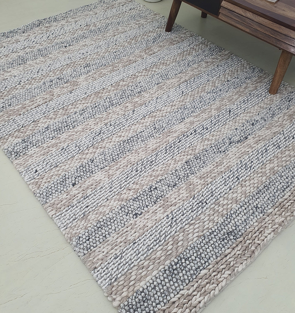 The RAMSHA HOME Wool Bedroom Rug, Living room Rug SR-050, a modern flatweave with chunky knit stripes in beige, gray, and white, lies on a light floor beside a wooden table leg.