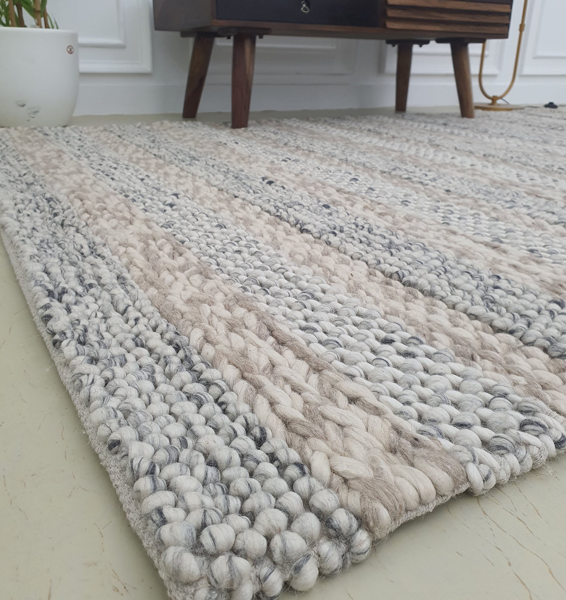 A close-up of the RAMSHA HOME Wool Bedroom Rug, Living room Rug, Floor Woven Rug SR-050 in textured beige and gray on a light floor, with a potted plant and wooden furniture visible against a white wall.