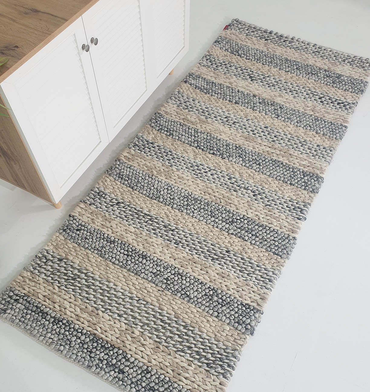 The RAMSHA HOME Wool Bedroom Rug, Living Room Rug SR-050 features alternating thick beige and gray stripes and is shown on a white floor beside a wooden cabinet with white doors.