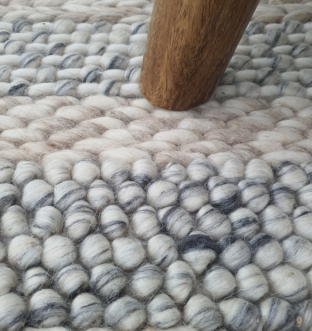 Close-up of a wooden furniture leg on the RAMSHA HOME Wool Bedroom Rug, Living Room Rug, Floor Woven Rug SR-050, featuring chunky woven stripes in white, gray, and cream fibers.