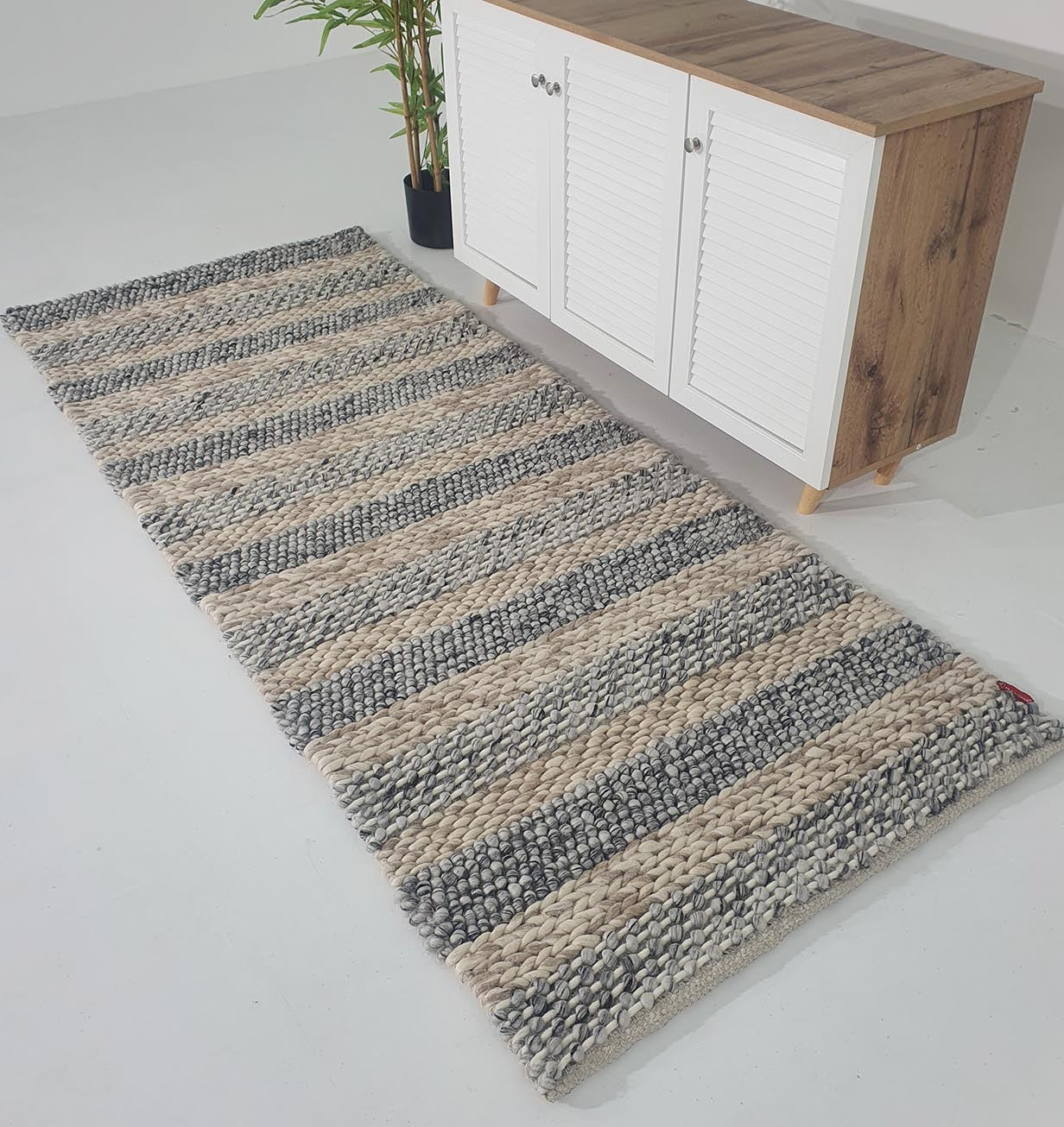 The RAMSHA HOME Wool Bedroom Rug SR-050, a modern flatweave with thick beige and gray stripes, is placed on a white floor beside a wooden cabinet with white doors and a potted green plant.