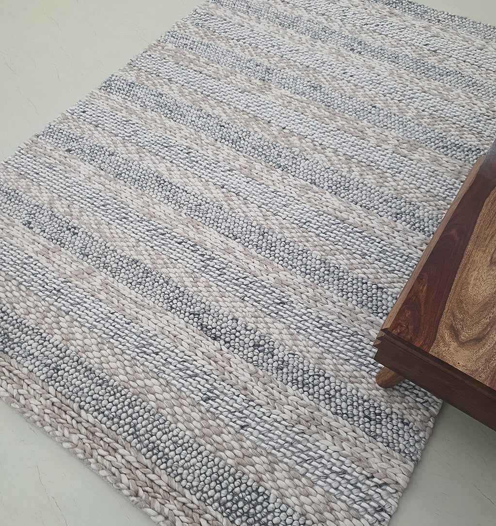 A close-up of the RAMSHA HOME Wool Bedroom Rug SR-050, a modern flatweave rug with light gray and beige stripes, shown on a light floor beside a wooden table. Perfect for living rooms or bedrooms.
