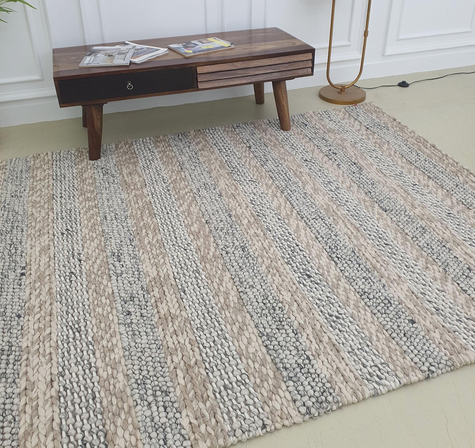 The RAMSHA HOME Wool Bedroom Rug, Living Room Rug, Floor Woven Rug SR-050 with beige and gray stripes is placed on a light floor beside a wooden coffee table with magazines, a gold floor lamp, and white paneled walls for a clean, modern look.