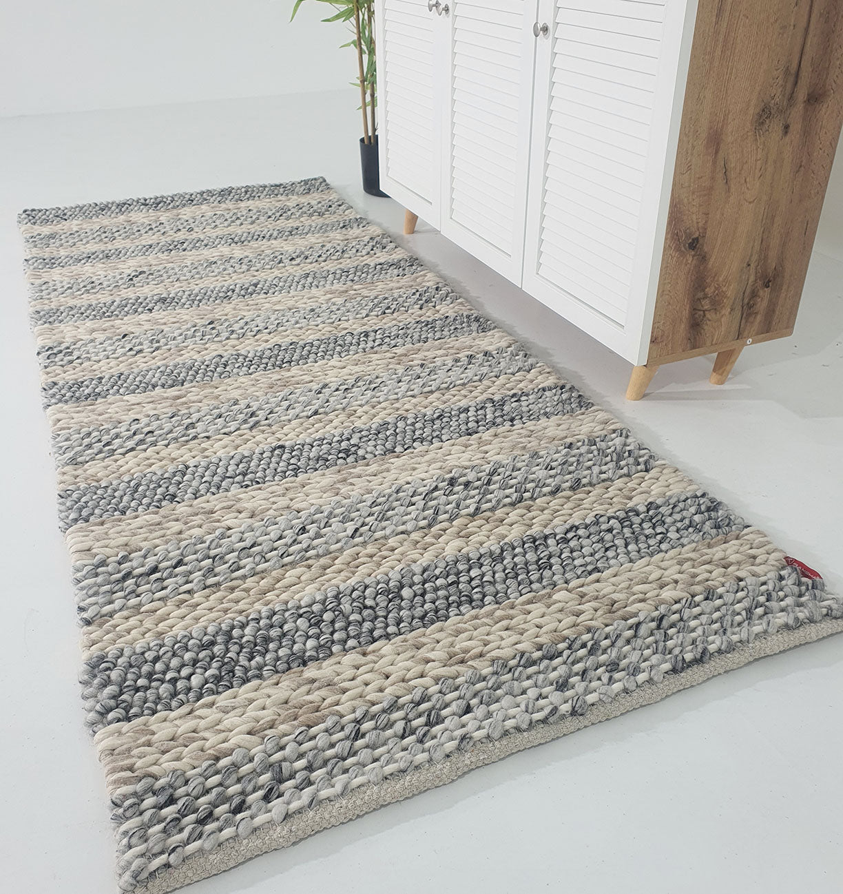 The RAMSHA HOME Wool Bedroom Rug (SR-050), a modern flatweave with thick gray and cream stripes, lies on a white floor beside a wooden cabinet with white doors and tapered legs; a green plant is visible in the background.