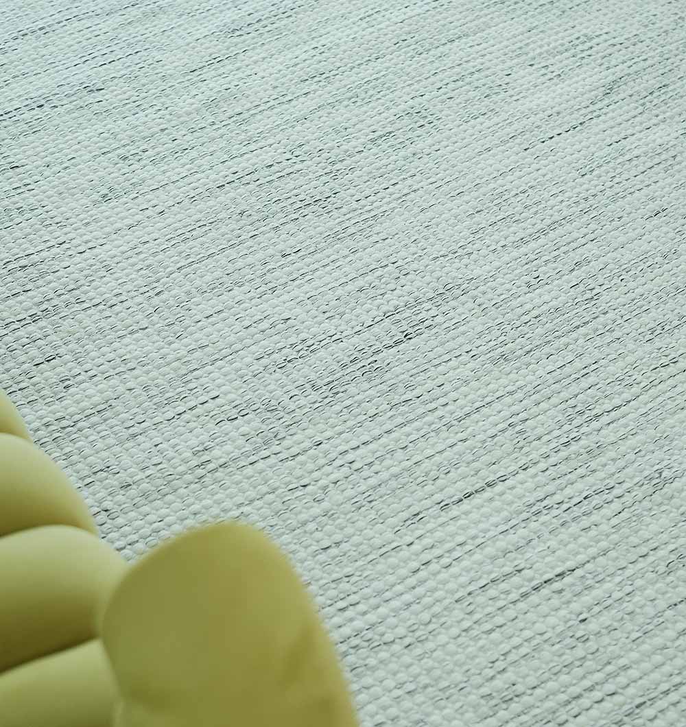 A close-up of the RAMSHA HOME Durable Handmade Natural White Woven Area Rug (SR-058) shows its subtle textured pattern, with the edge of a pale yellow curved piece of furniture visible in the lower left corner.
