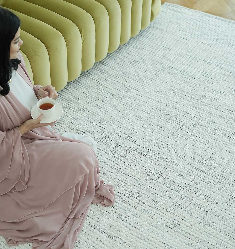 A woman in a pale pink robe sits on the RAMSHA HOME Durable Handmade Natural White Woven Area Rug SR-058, holding a cup of tea. She is near a green tufted sofa, with only her upper body partially visible in the image.