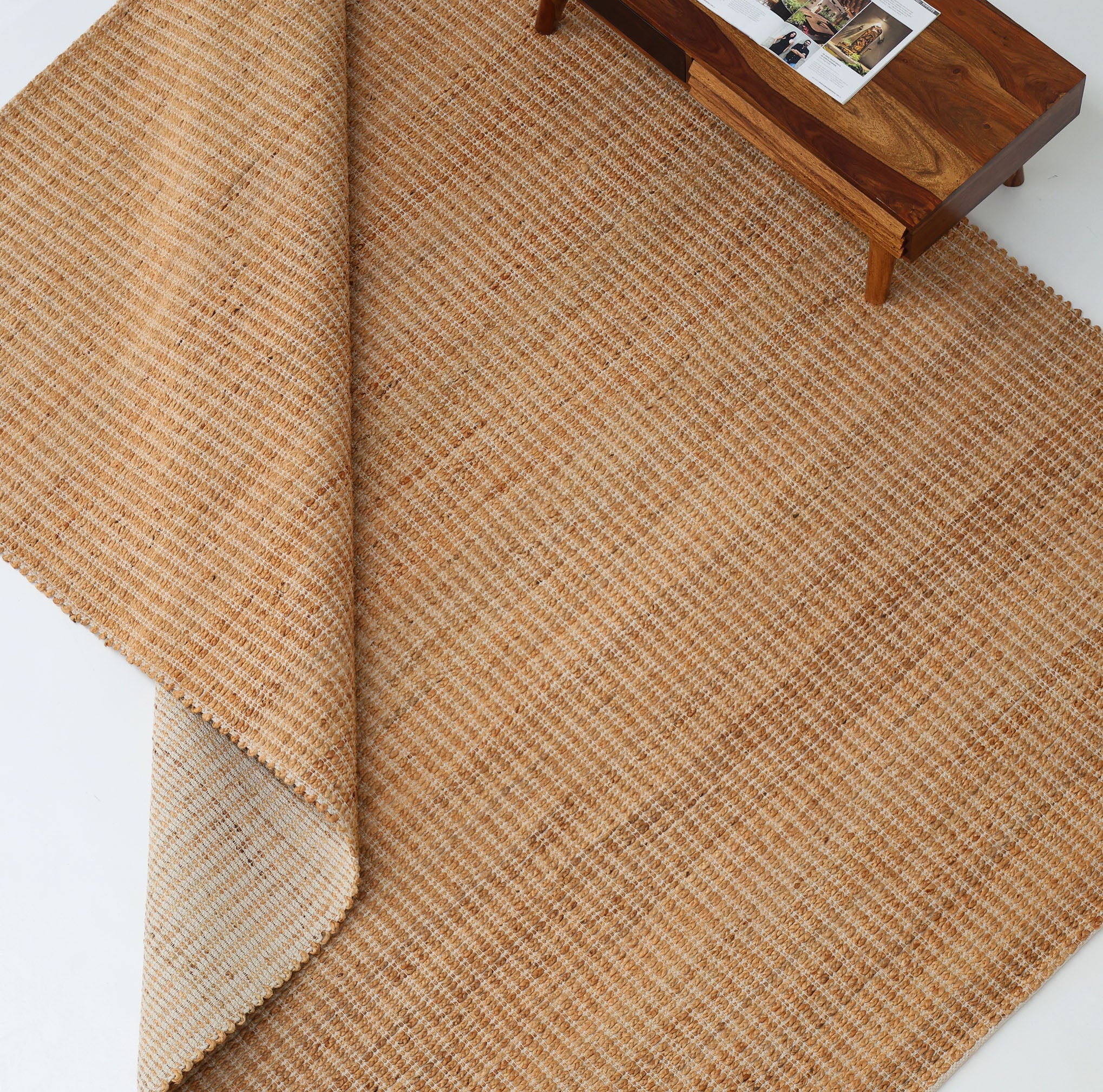 The RAMSHA HOME Handmade Natural Color Hand Woven Area Rug SR-062 is partially folded on a white floor beside a wooden table with framed photos and a magazine, bringing artisanal charm to the décor.