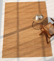 Sunlight streams over the RAMSHA HOME Handmade Natural Color Hand Woven Area Rug SR-063. A wooden chair with a cane seat and a round side table holding a teapot and two cups rest on this artisanal home décor piece.
