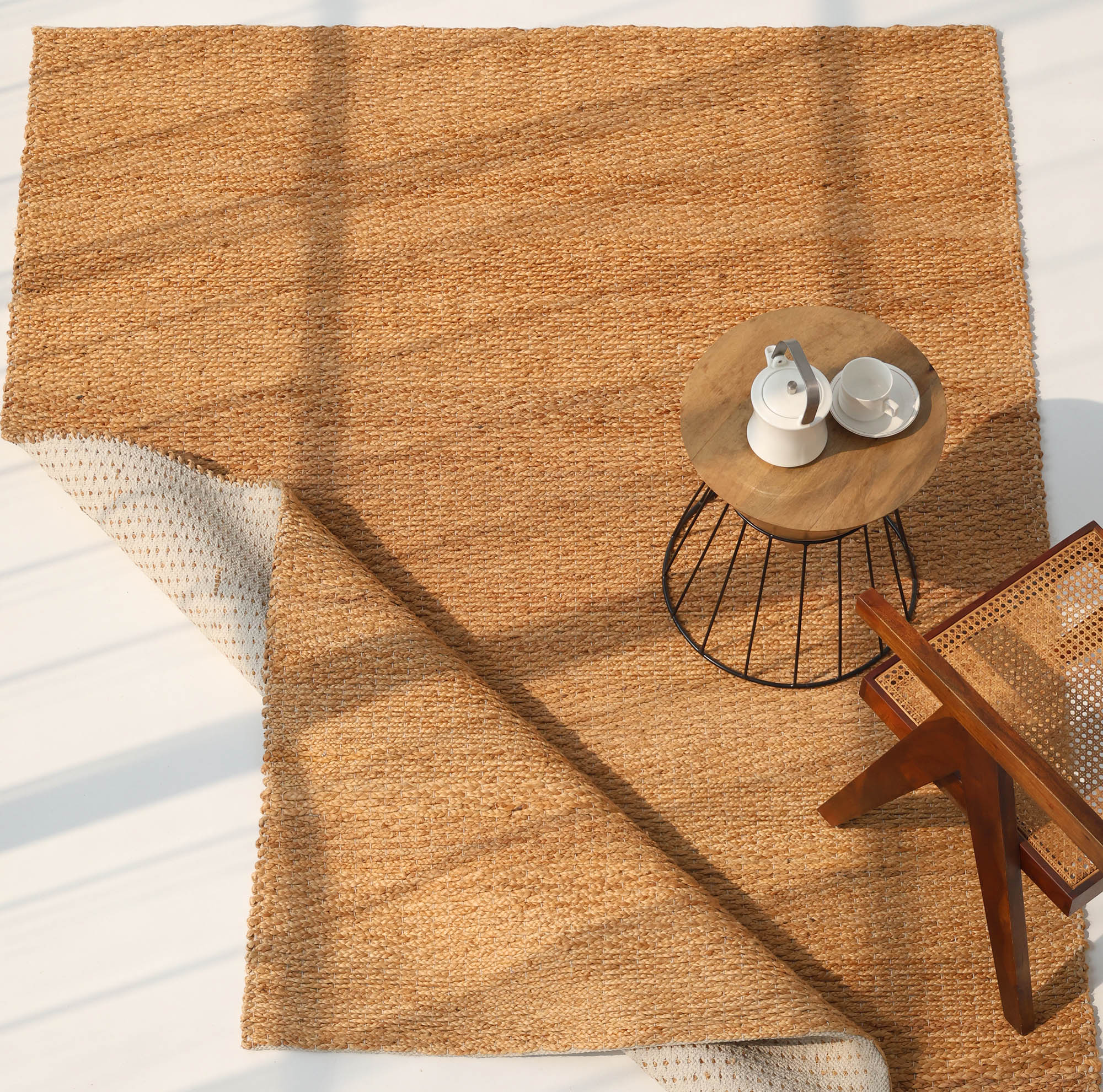 The Handmade Natural Color Hand Woven Area Rug SR-063 by RAMSHA HOME lies on the floor with a corner flipped over. A round side table with a teapot and cups sits nearby, next to a cane-seated chair, all in natural sunlight.