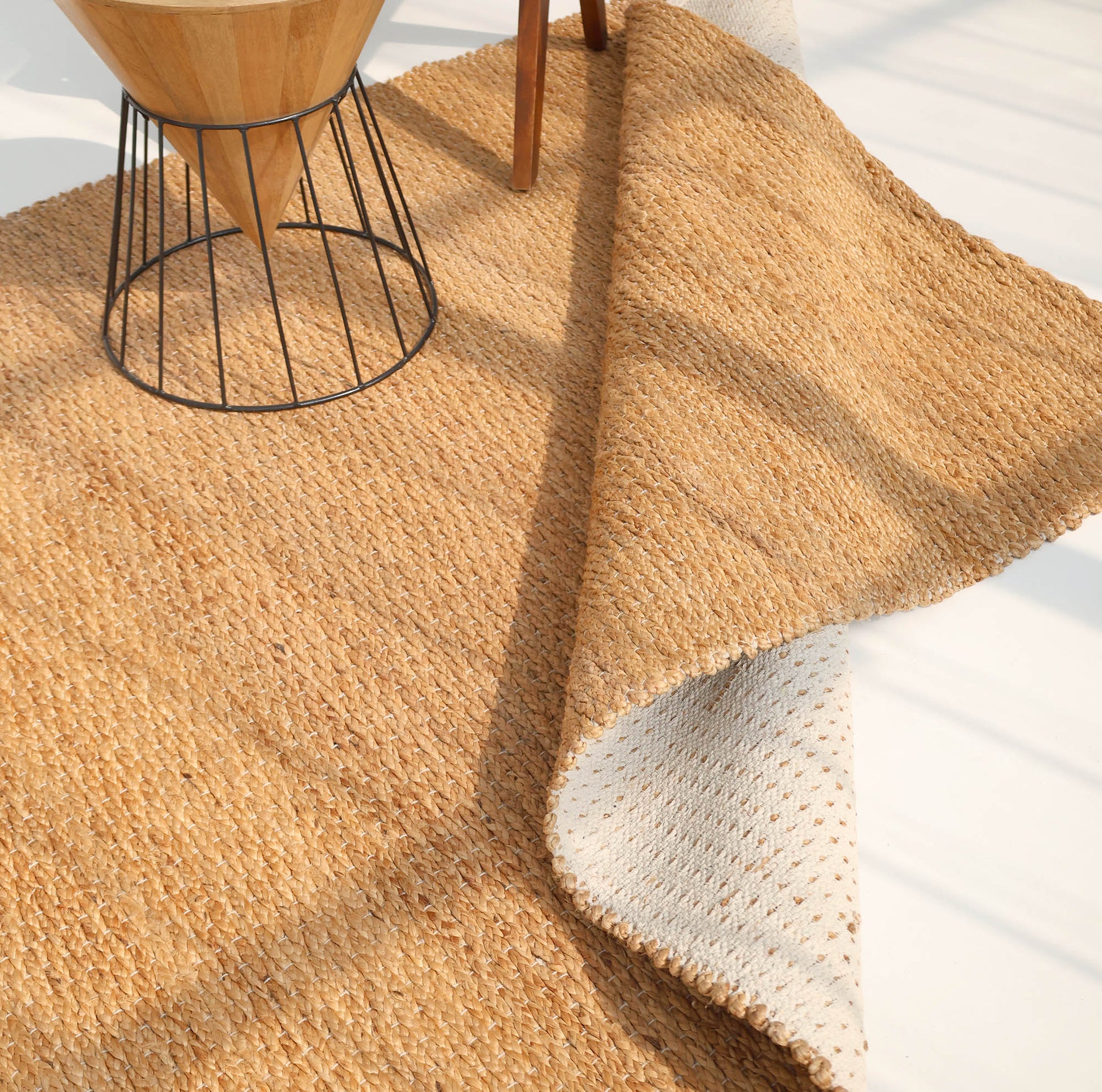 The RAMSHA HOME Handmade Natural Color Hand Woven Area Rug SR-063 is partially flipped, showing its lighter underside, with the corner of a wooden table with metal legs resting on it over a light-colored floor.