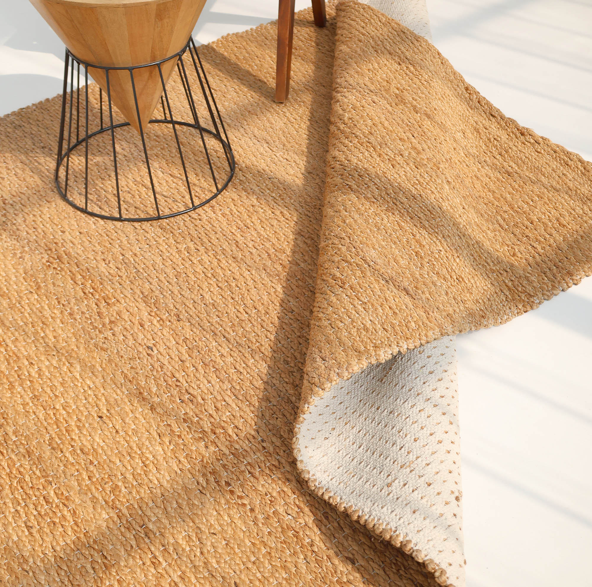 The RAMSHA HOME Handmade Natural Color Hand Woven Area Rug SR-063 is partially flipped, showing its lighter underside, with the corner of a wooden table with metal legs resting on it over a light-colored floor.